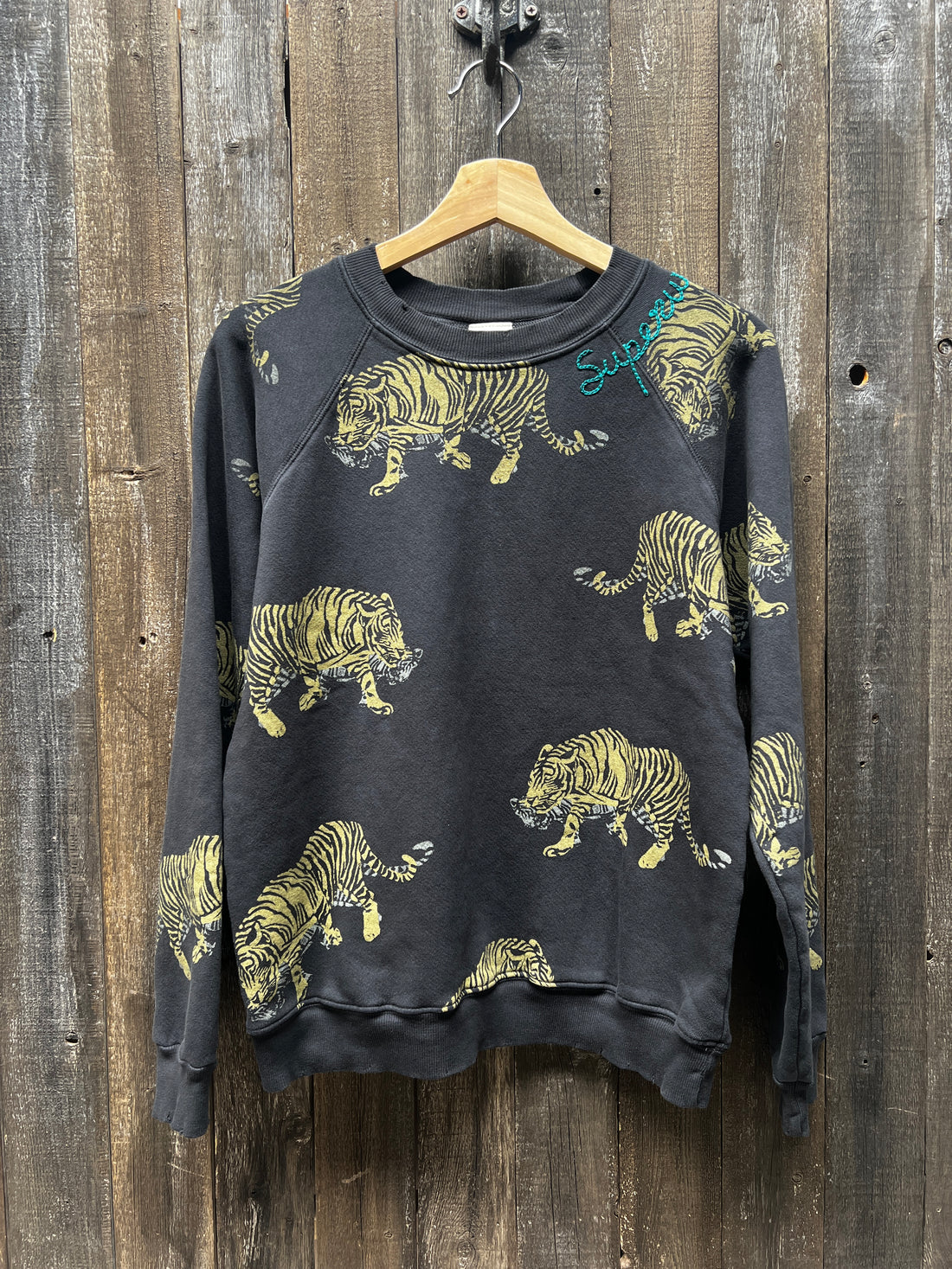 A "C&S Sweats Into the Wild Superwoman" sweatshirt from I Stole My Boyfriend's Shirt, featuring a silk screen print of yellow and black tigers, is hanging on a wooden wall. The teal "Supreme" logo on the upper left side near the shoulder adds a striking contrast to this bold design.