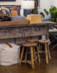 A rustic wooden table with four drawers, adorned with a basket, a lamp, and decorative items sits in the space. Underneath are three imported ROUND STOOLs by Indus Design Imports that add a handmade vintage charm. The area is also decorated with a white pouf and various plants. In the background, pillows and furniture can be seen, enhancing the cozy atmosphere with timeless elegance.