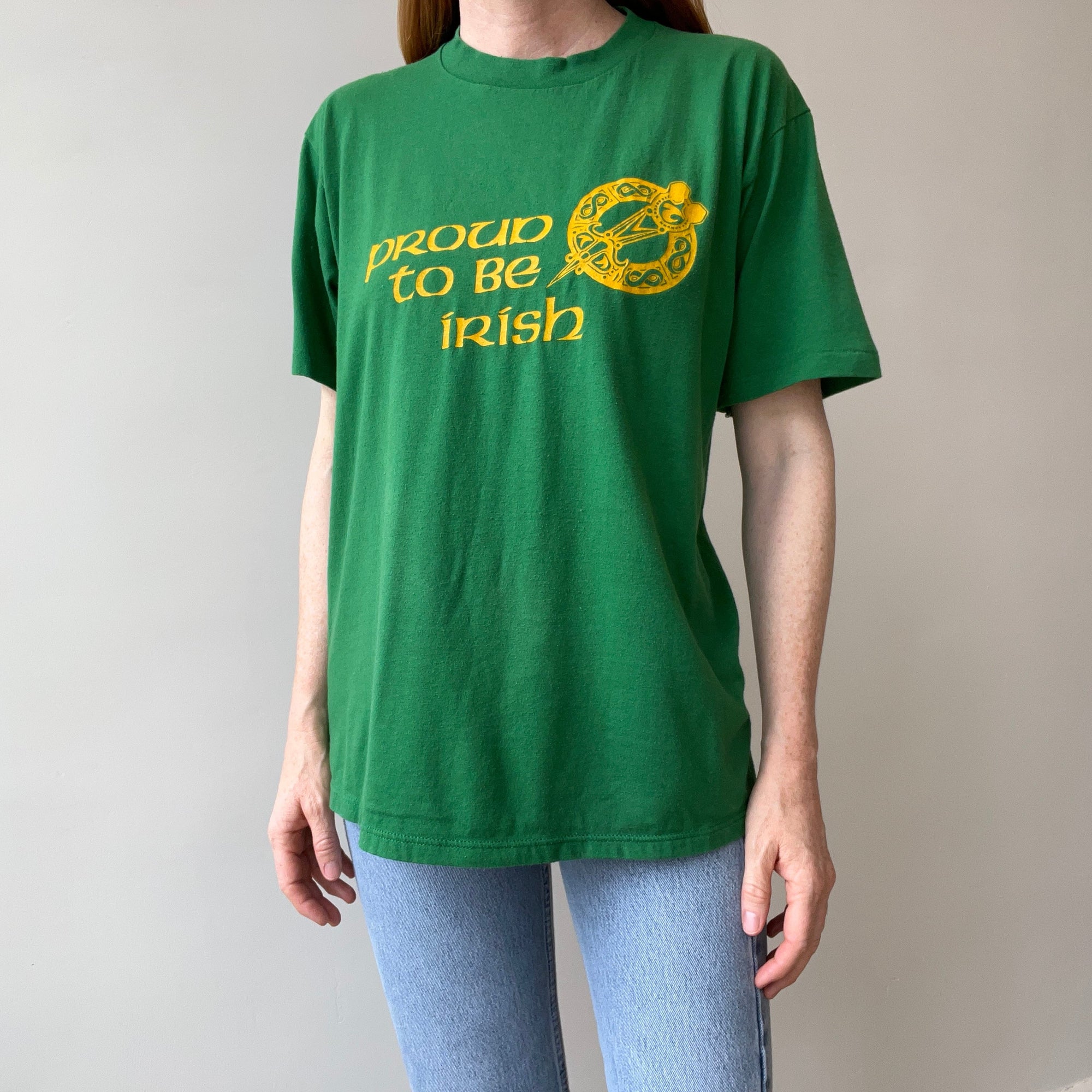 A tall person in a vintage green "80s Proud to Be Irish Tee" by Red Vintage Co., size XL, with yellow text reading "Proud to be Irish," stands against a plain background. The shirt features a decorative circular design and pairs perfectly with light blue jeans.