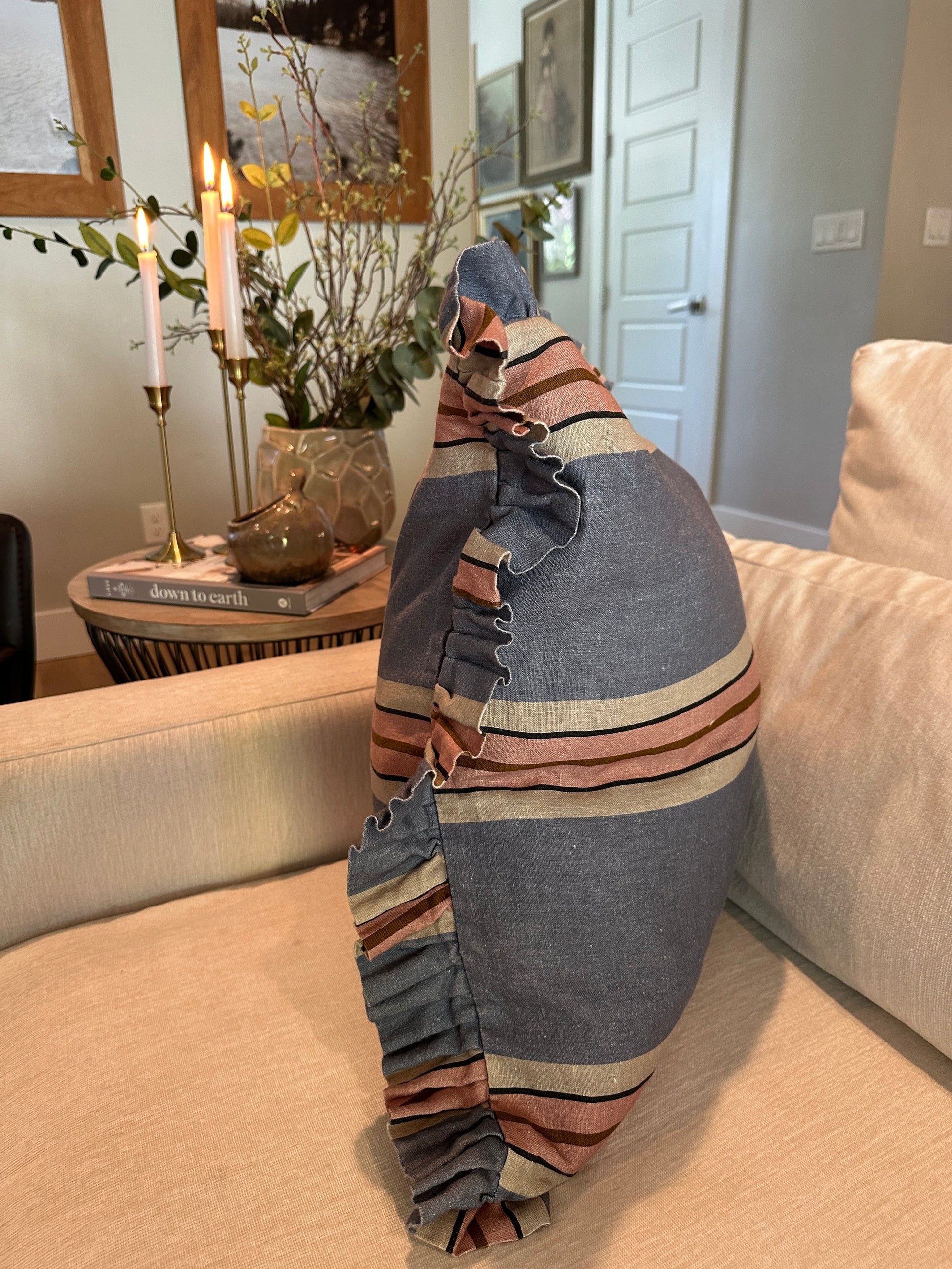 The Rennah Collective Foxy Blue Striped Ruffle Pillow 22x22 sits on a beige sofa in a cozy living room, surrounded by candles, books, flowers, and framed pictures for a welcoming touch.