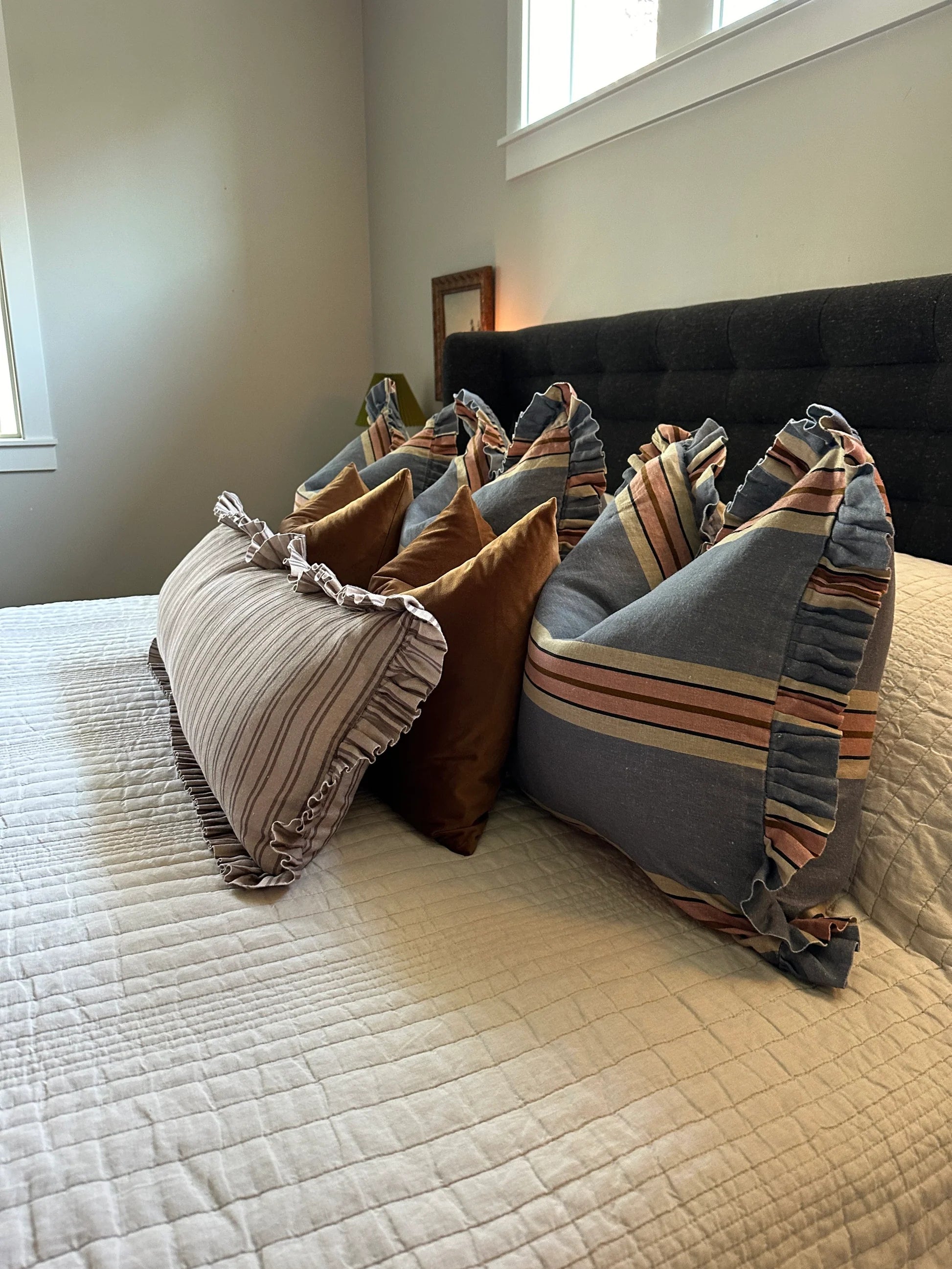 A neatly made bed with a dark upholstered headboard features four decorative pillows: two brown velvet and two Rennah Collective Foxy Blue Striped Ruffle Pillows (22x22), all arranged on a crisp white quilted bedspread.