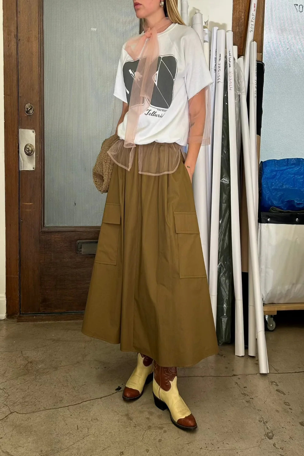 Indoors, a person models the Aquarius Cocktail Le Cargo Skirt with large pockets, paired with a graphic tee featuring sheer pink at the neckline, cream and brown cowboy boots, and a woven bag. Rolls of paper appear in the background.