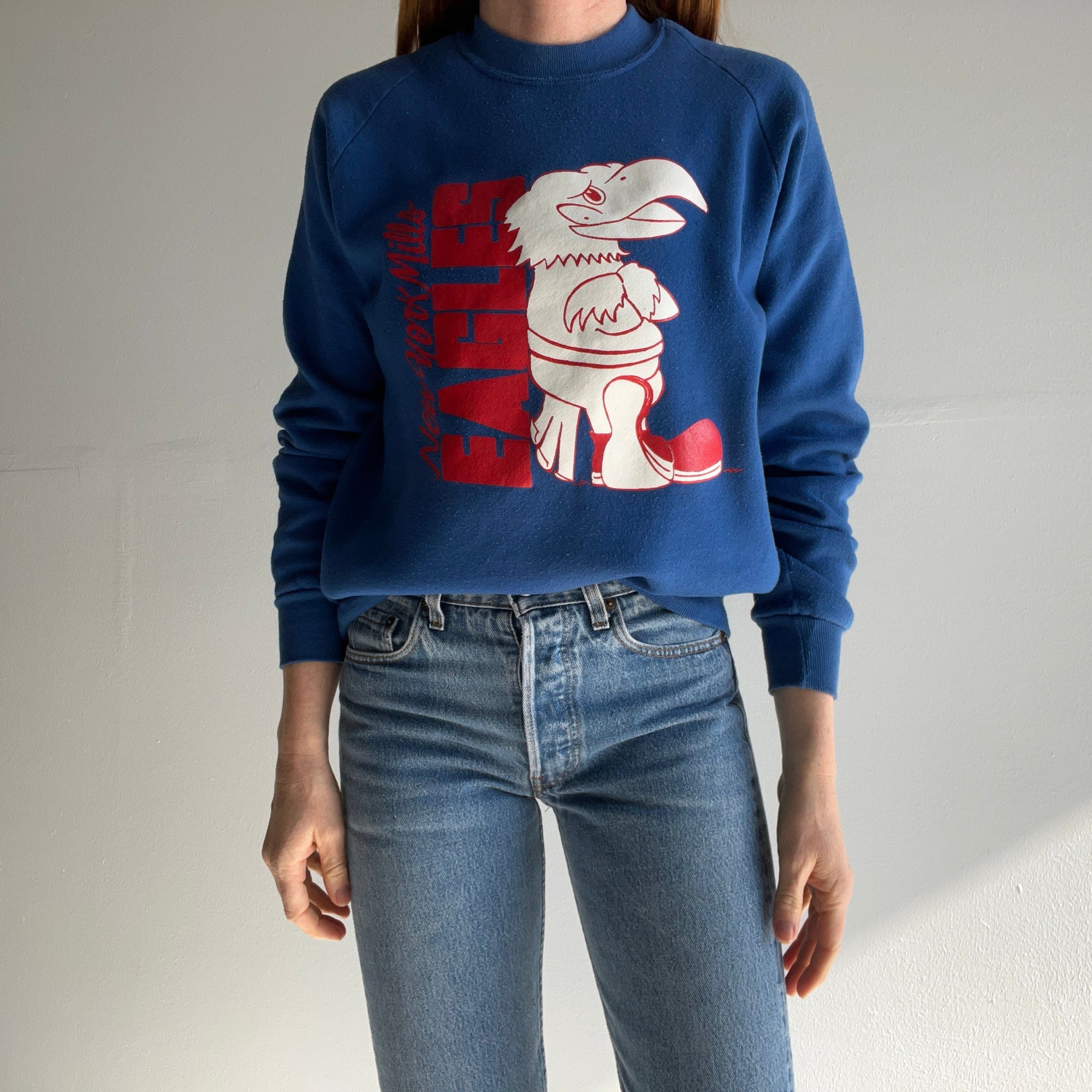 A person wearing the Red Vintage Co.'s 80s New York Mills Eagles Sweatshirt, featuring a large red and white cartoon eagle on vintage blue, stands against a plain wall. Combined with light blue jeans, the outfit radiates a soft and cozy vibe characteristic of the New York Mills aesthetic.