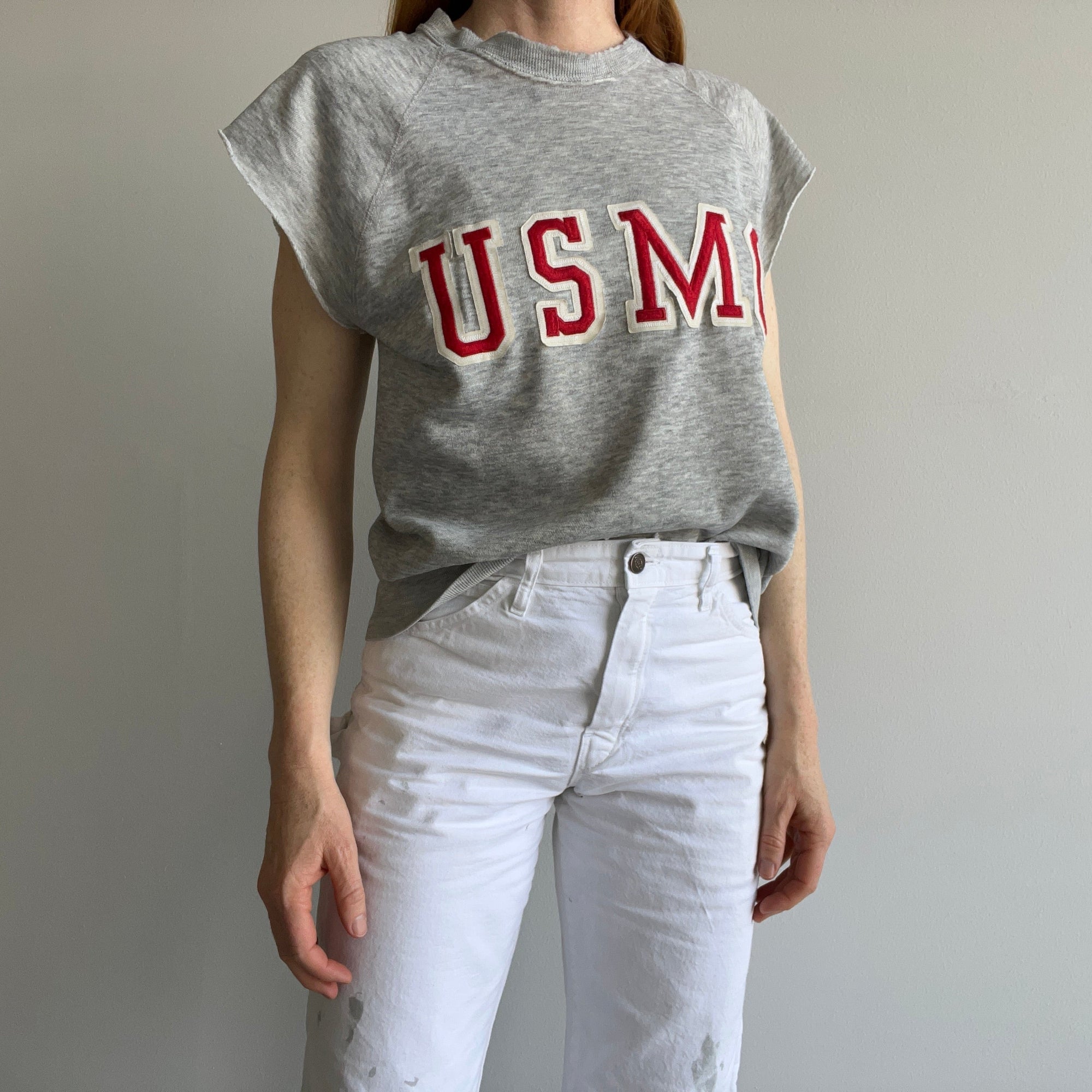 A person wearing an 80s USMC Short Sleeve Sweatshirt by Red Vintage Co., paired with white pants, stands against a plain light-colored wall.