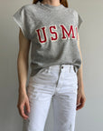 A person wearing an 80s USMC Short Sleeve Sweatshirt by Red Vintage Co., paired with white pants, stands against a plain light-colored wall.