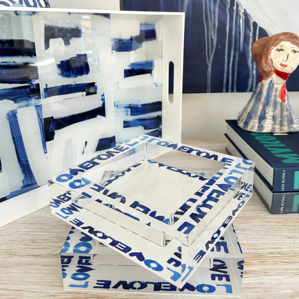 A Love on Repeat deco bowl with abstract blue and white architectural designs in Bungalow style is opened on a wooden surface, with a painted figurine of a girl standing to the right, creating an artistic display by Kerri Rosenthal.