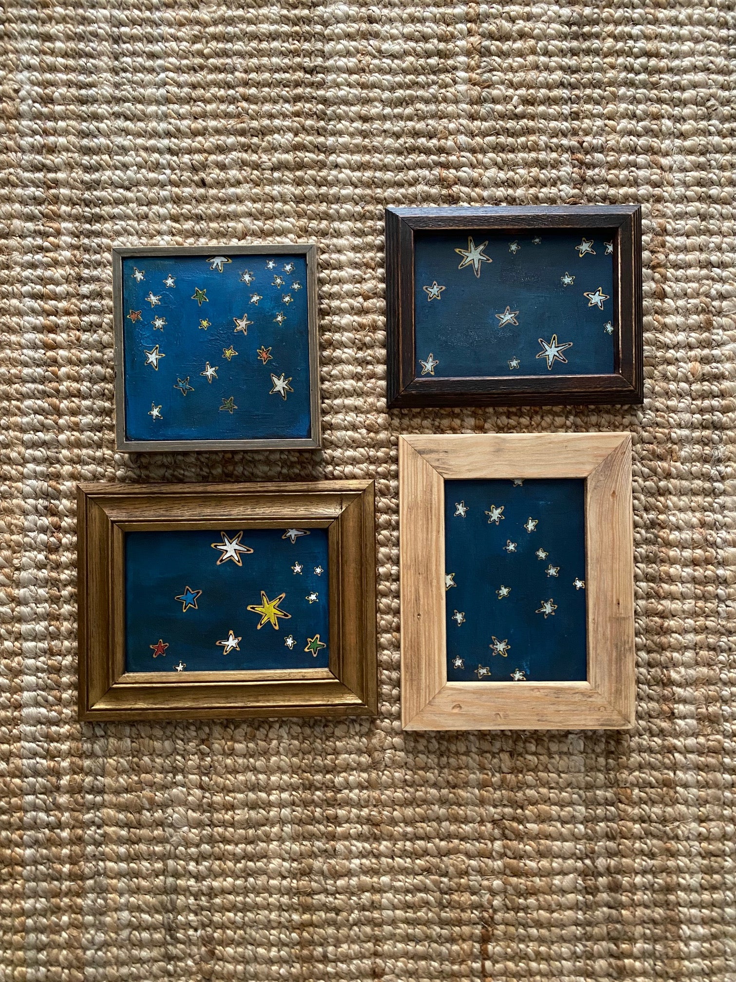SKY & STARS by Tak Design features four hand-painted artworks with gold stars on dark blue, each in a unique vintage wood frame. Arranged in a rectangular grid on a textured woven surface, the set adds cosmic charm to any space.