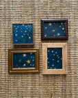 SKY & STARS by Tak Design features four hand-painted artworks with gold stars on dark blue, each in a unique vintage wood frame. Arranged in a rectangular grid on a textured woven surface, the set adds cosmic charm to any space.