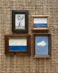 Four small framed artworks from Tak Design's SKY & STARS: hand-painted pieces by a local artist, featuring scissors, two minimalist blue seascapes, and a white cloud in a blue sky on a woven textured background.