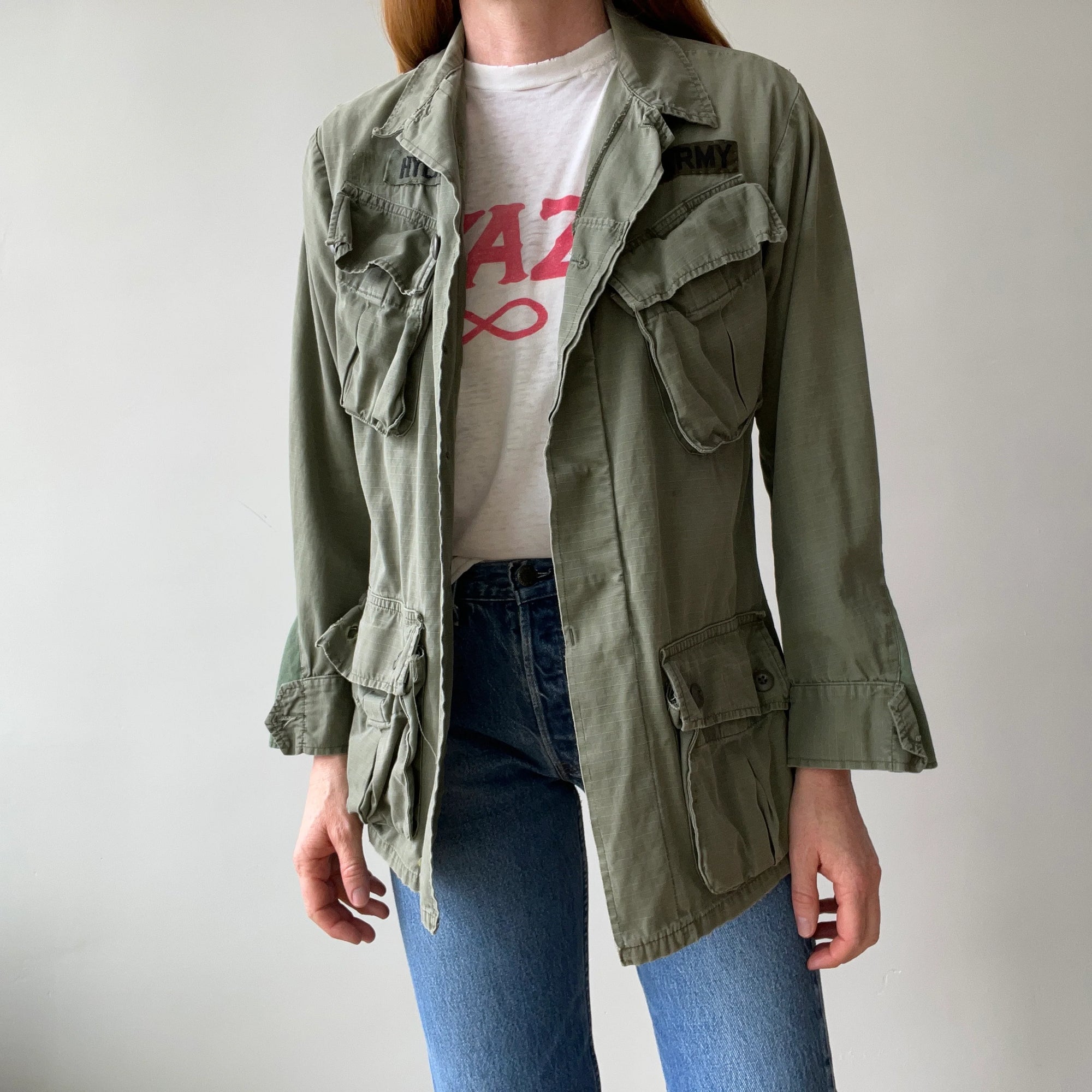 A person wearing a Red Vintage Co. 1969 Cotton Jungle Jacket in light green over a white graphic T-shirt and blue jeans, standing against a plain light background.