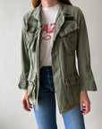 A person wearing a Red Vintage Co. 1969 Cotton Jungle Jacket in light green over a white graphic T-shirt and blue jeans, standing against a plain light background.