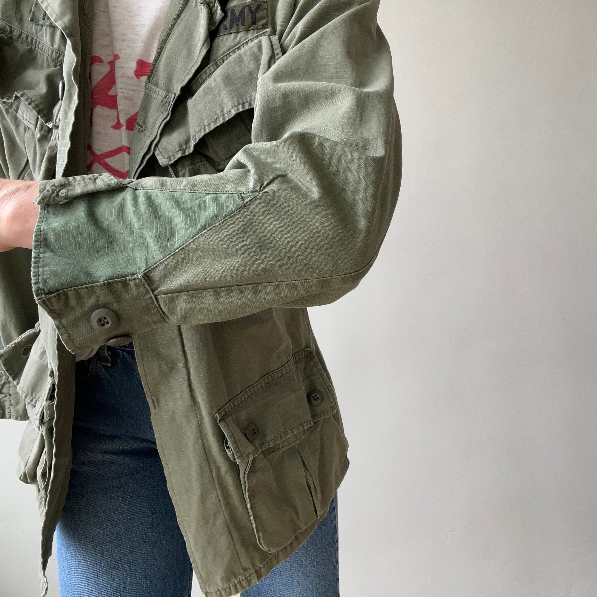 A person in blue jeans adjusts the sleeve of a 1969 Cotton Jungle Jacket by Red Vintage Co., featuring &quot;ARMY&quot; lettering. The plain, light background adds an East Hollywood vibe.