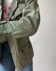 A person in blue jeans adjusts the sleeve of a 1969 Cotton Jungle Jacket by Red Vintage Co., featuring "ARMY" lettering. The plain, light background adds an East Hollywood vibe.