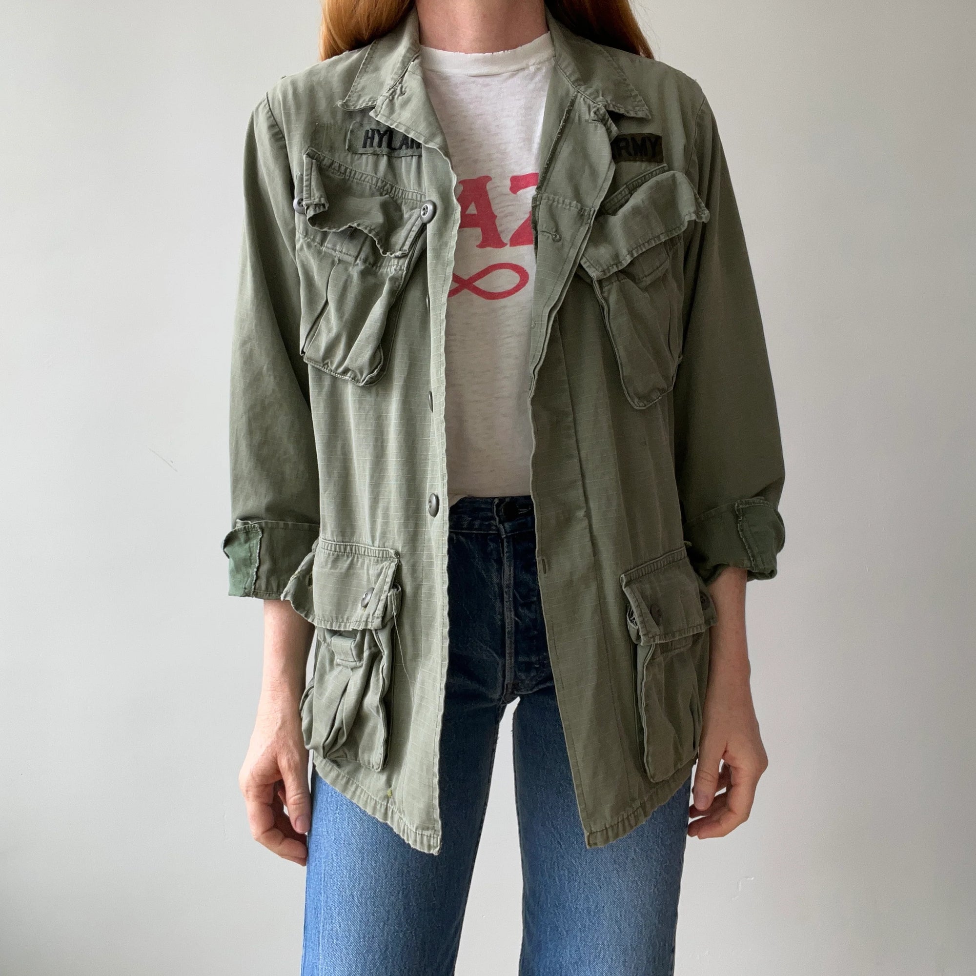 Someone with long hair wears the Red Vintage Co. 1969 Cotton Jungle Jacket in light green, featuring multiple pockets and black name patches, over a white tee with red lettering and blue jeans against a plain, light background.
