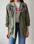 Someone with long hair wears the Red Vintage Co. 1969 Cotton Jungle Jacket in light green, featuring multiple pockets and black name patches, over a white tee with red lettering and blue jeans against a plain, light background.