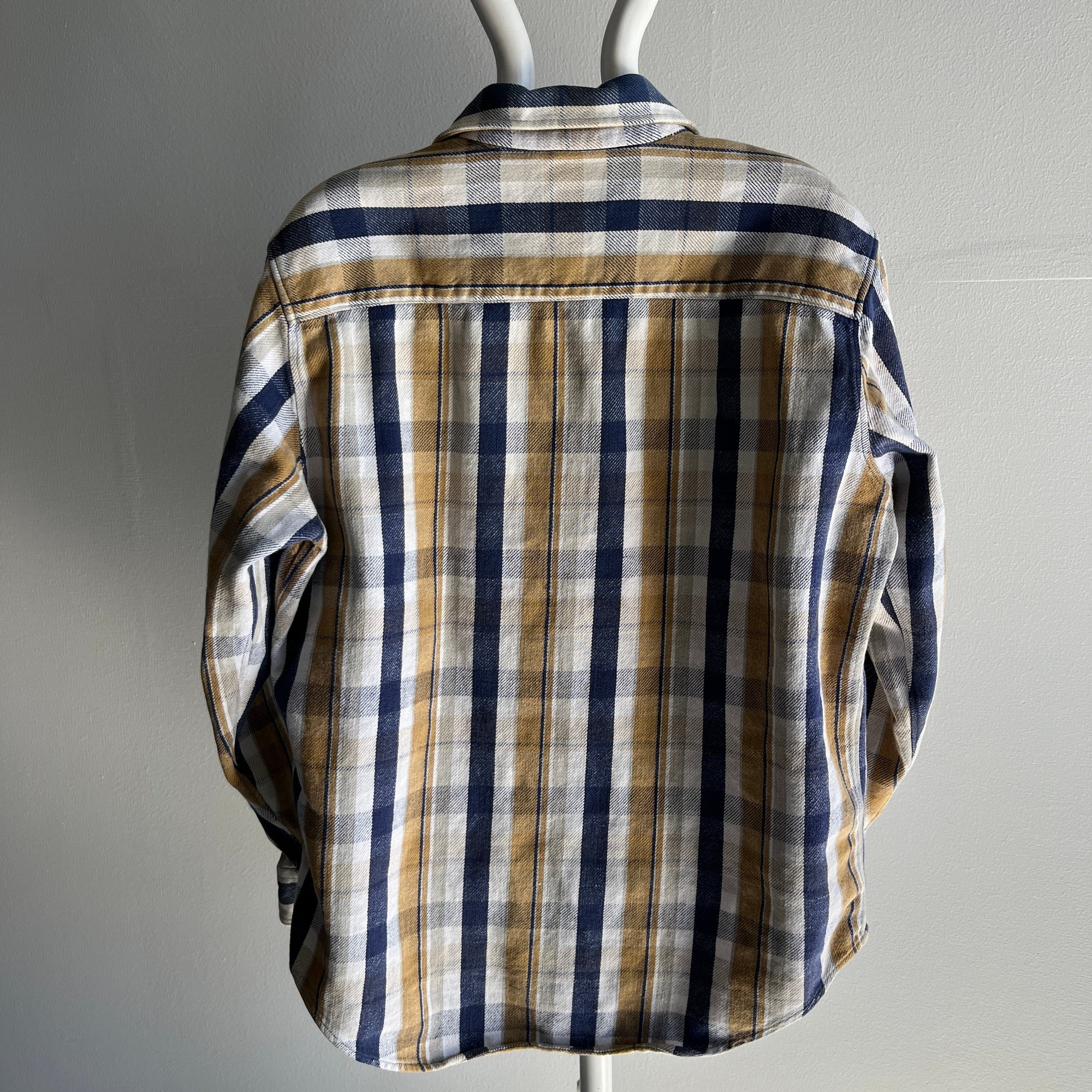 The 90/2000s Neutrals Navy Flannel by Red Vintage Co. is showcased on a mannequin, featuring its vintage-grade appeal and long sleeves. The shirt's beige, white, and blue checks add charm, complemented by chest pockets, against a plain white wall backdrop.