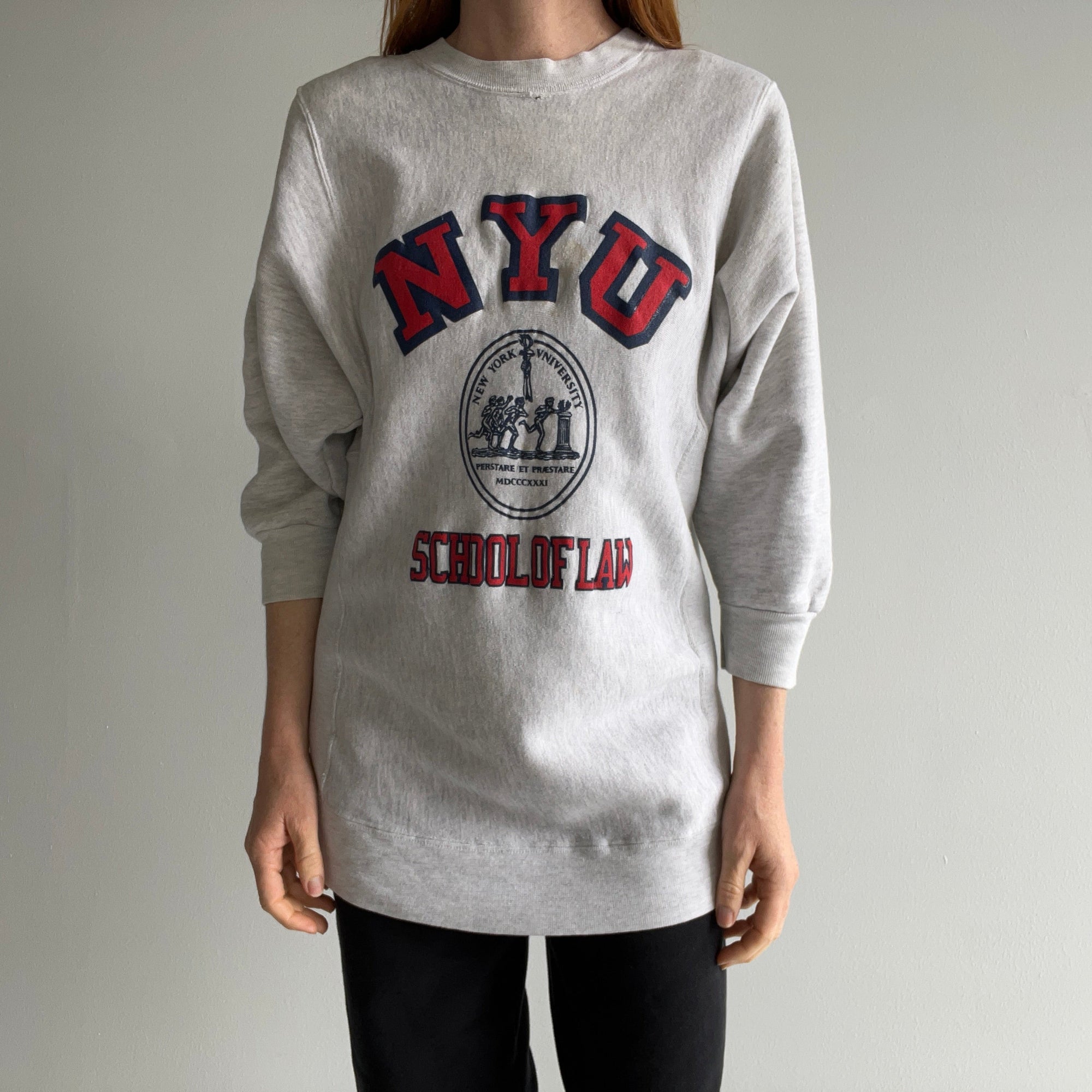 Dressed in a gray 80s NYU Law School Half Sleeve Sweatshirt by Red Vintage Co., which showcases "NYU School of Law" and a vintage circular seal graphic, a person stands against a plain background, paired with black pants.