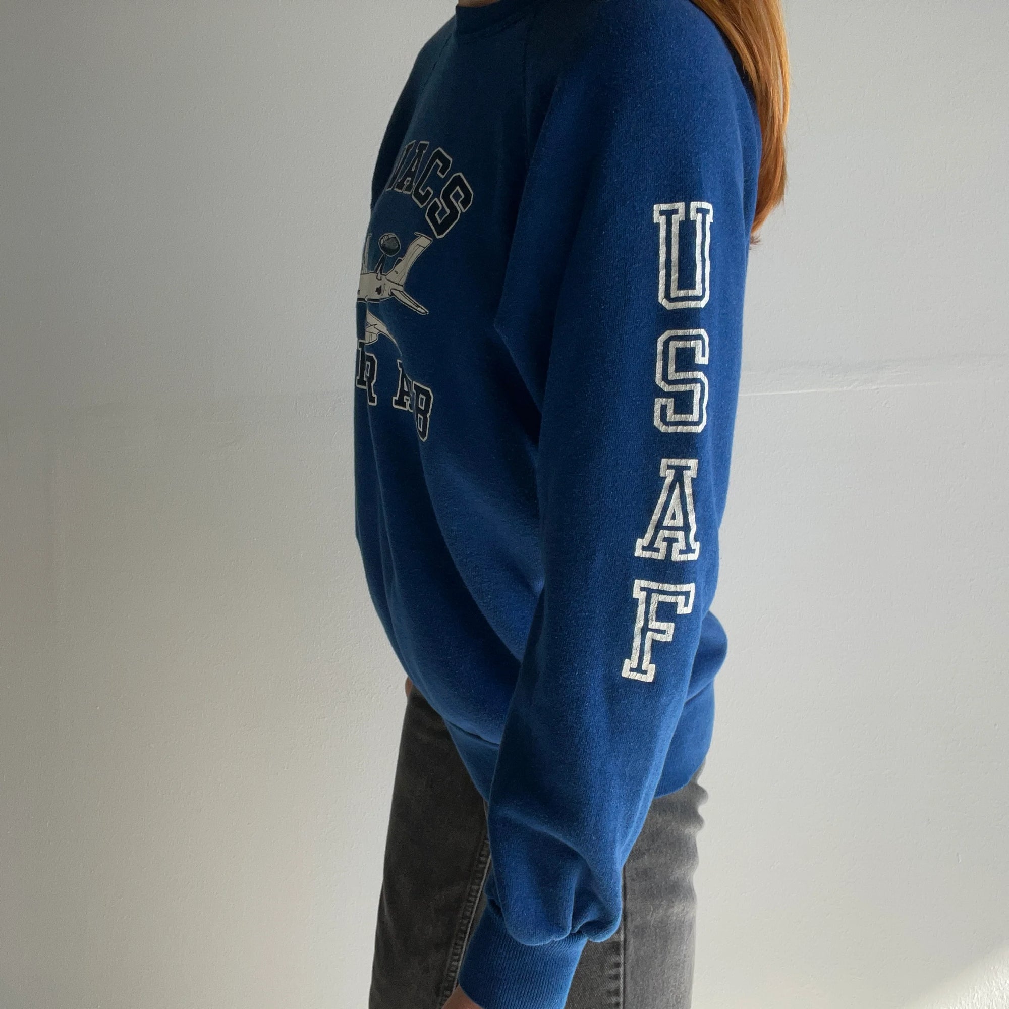 Clad in an 80s AWACS Tinker Air Force Base Sweatshirt from Red Vintage Co., a person stands against a plain white background. The sleeve features the letters "USAF," and the chest bears a circular emblem showcasing a bird in flight. The fabric's super soft texture suggests its lightly worn charm.