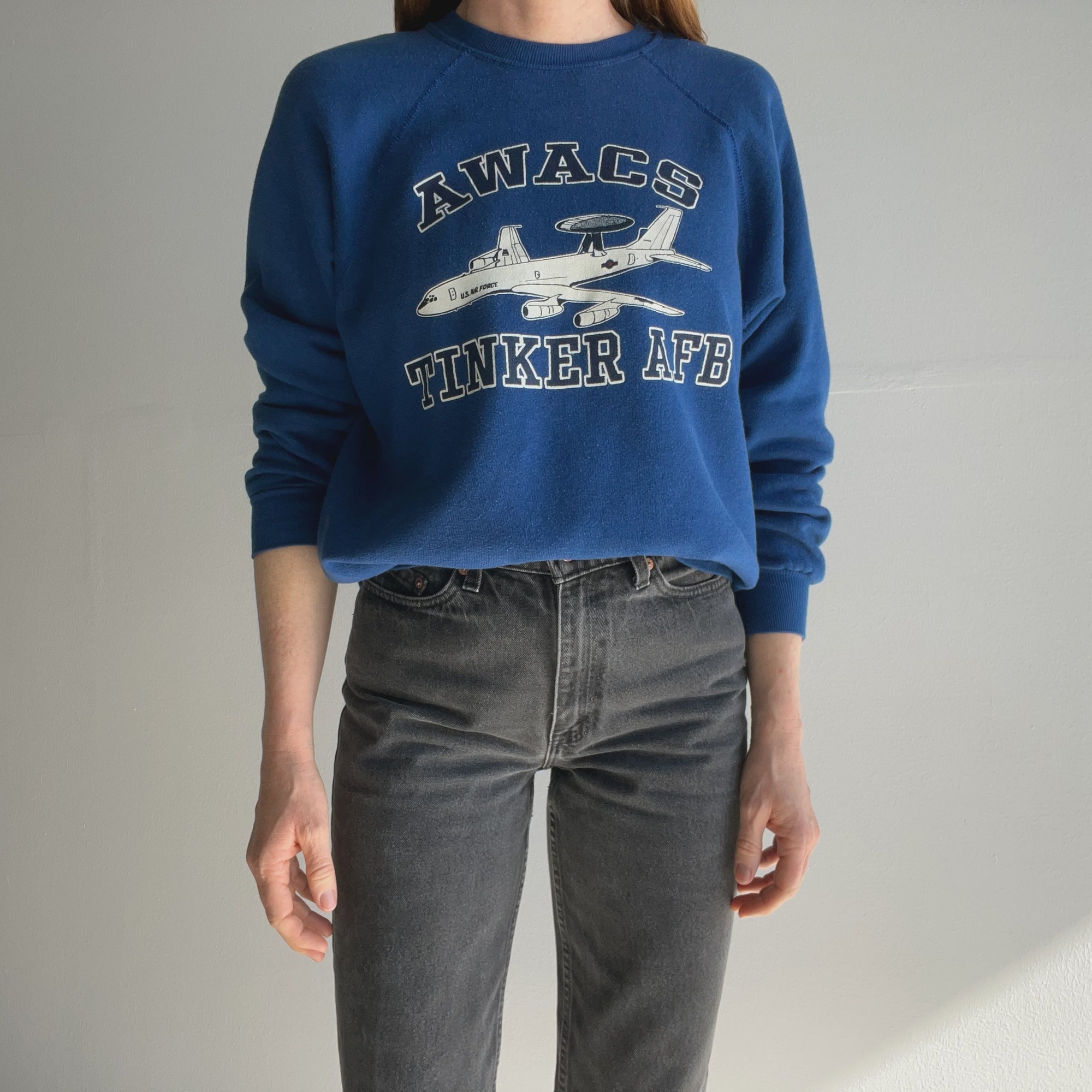 A person wearing a vintage "80s AWACS Tinker Air Force Base" sweatshirt by Red Vintage Co., featuring "AWACS Tinker AFB" and an airplane graphic, pairs it with dark jeans against a plain background. The ultra-soft fabric provides comfort and lends a gently worn charm to the look.