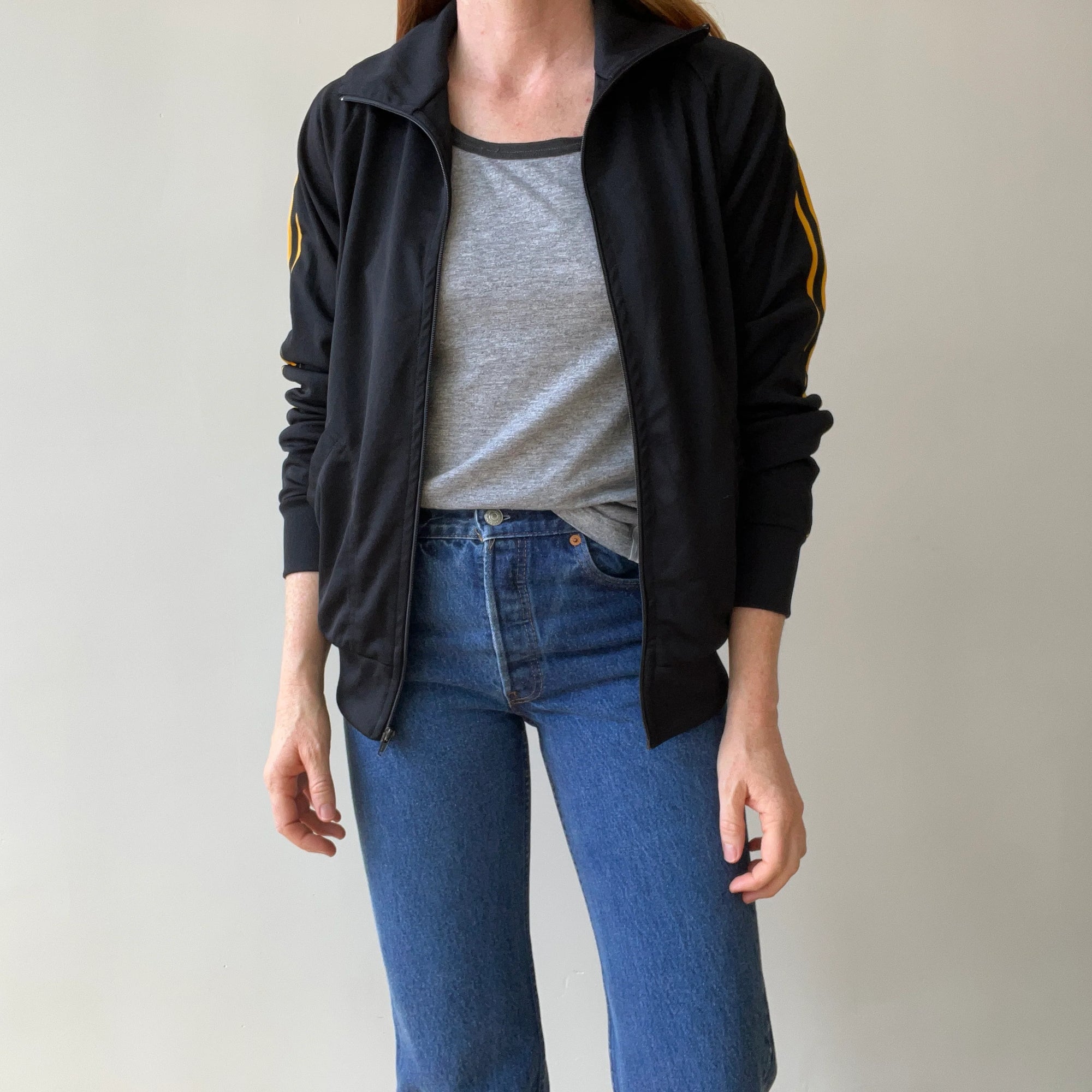 A person (head not visible) stands against a plain background wearing a lightly worn Red Vintage Co. 1980s Black & Yellow Track Jacket with yellow sleeve stripes and subtle shoulder drop, paired with a gray t-shirt and blue jeans.