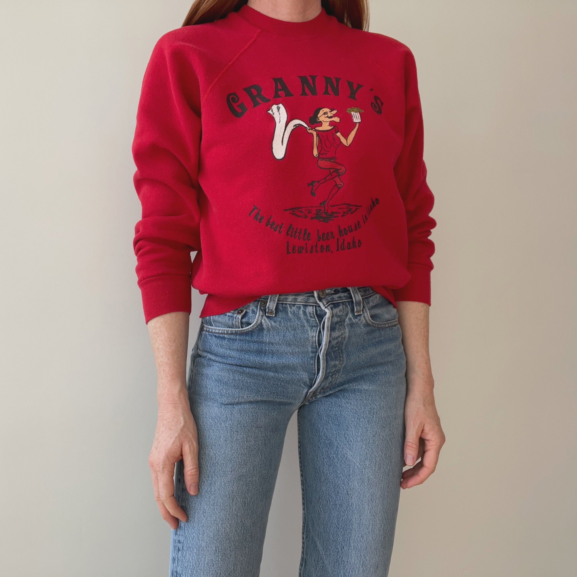 A person wearing an 80s The Best Little Beer House In Idaho sweatshirt by Red Vintage Co., featuring a graphic design of someone dancing and the text "Granny's - The best little beer house in Lewiston, Idaho," paired with blue jeans.