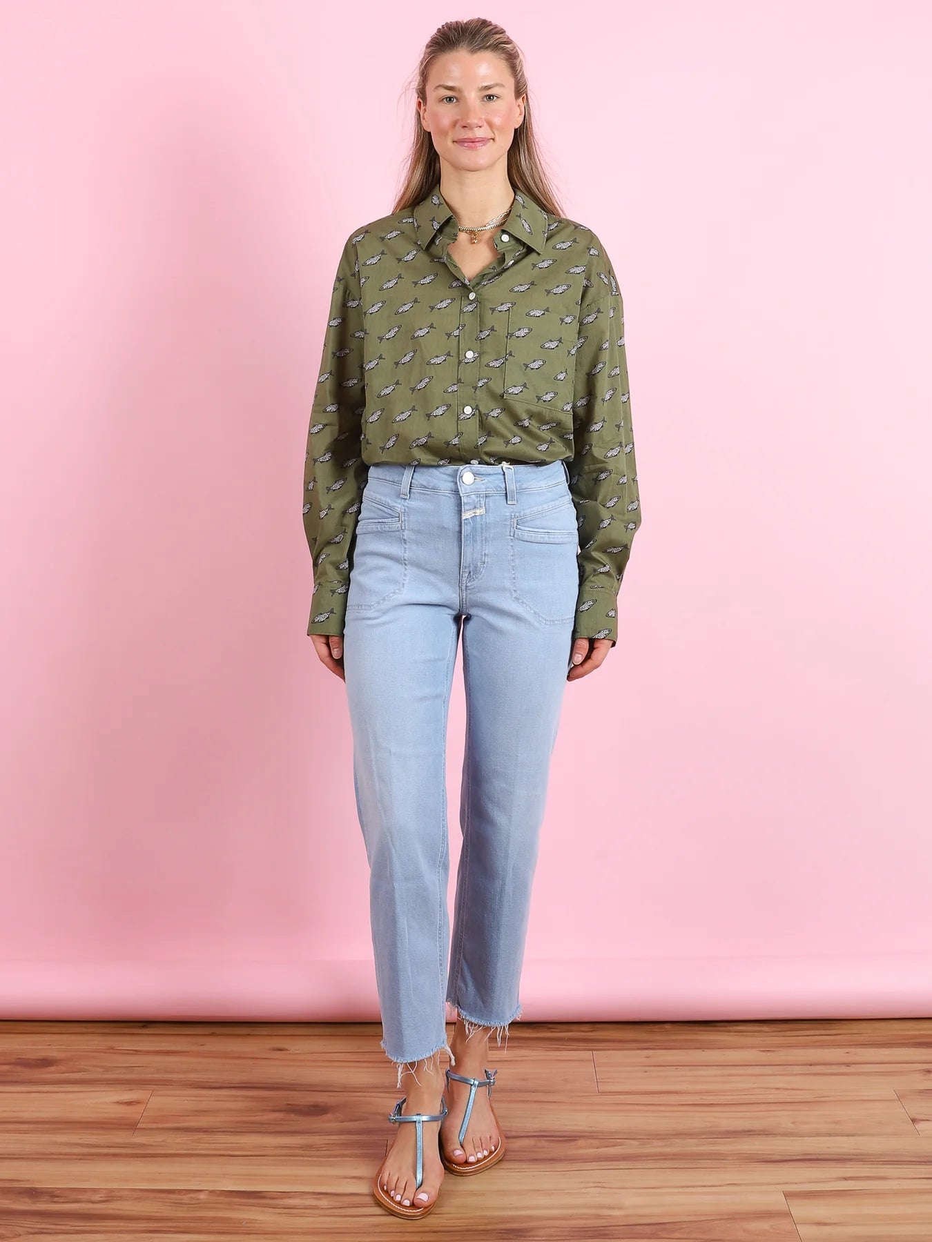 A woman with long, straight hair smiles at the camera, wearing a CLOSED Oversized Shirt against a pink background, paired with light blue high-waisted jeans featuring frayed hems and embroidered patches, and blue sandals.