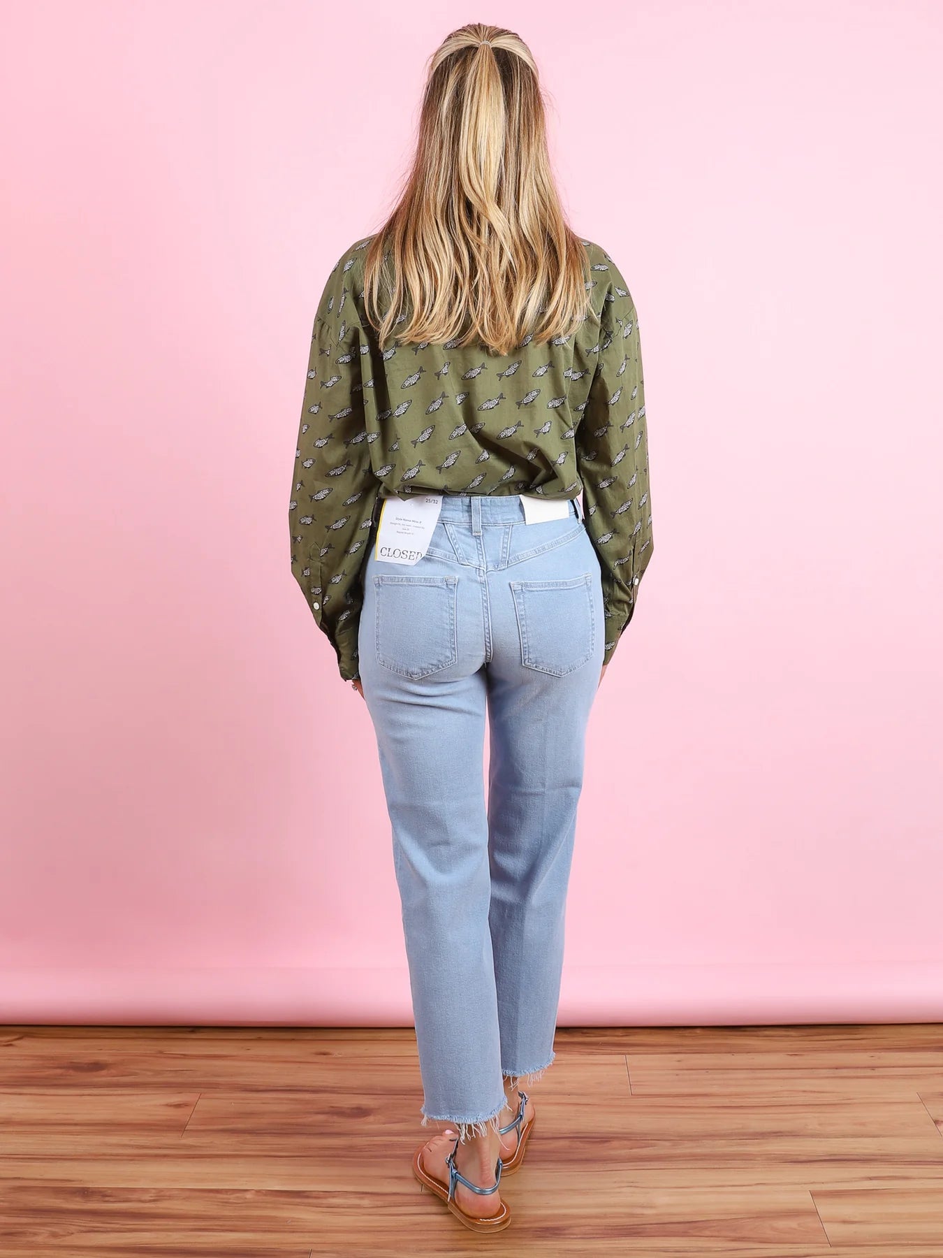 A woman with long blonde hair stands facing a pink wall, wearing the CLOSED Oversized Shirt, light blue jeans featuring a tag on the back pocket, sandals, and standing on a wooden floor.