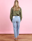 A woman with long blonde hair stands facing a pink wall, wearing the CLOSED Oversized Shirt, light blue jeans featuring a tag on the back pocket, sandals, and standing on a wooden floor.