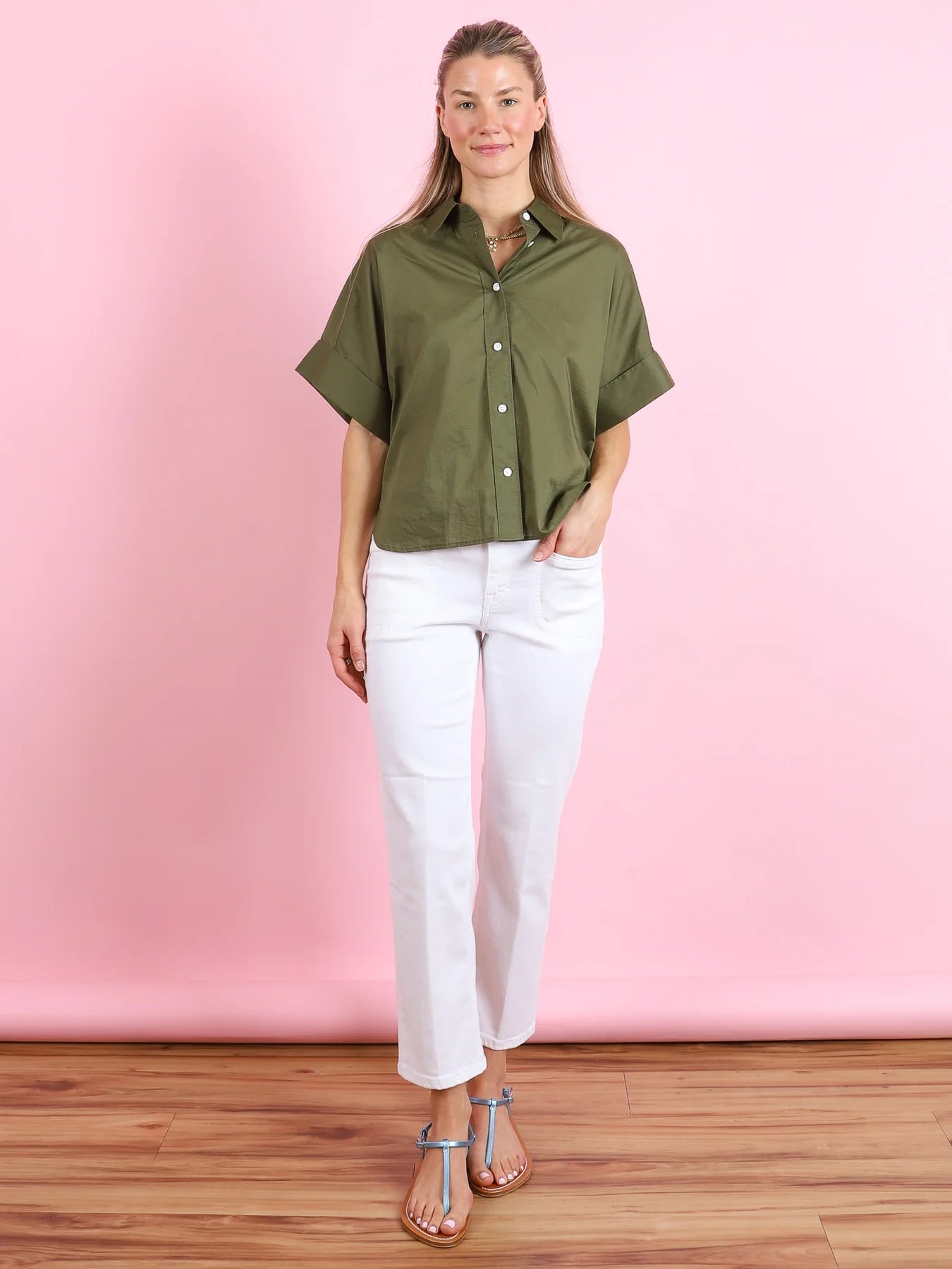 A woman with long, light brown hair stands against a pink background, smiling softly with one hand in her pocket. She wears CLOSED’s Turn-Up Shirt—a relaxed fit cotton shirt with a pointed collar and mother-of-pearl buttons—white jeans, and flat sandals.