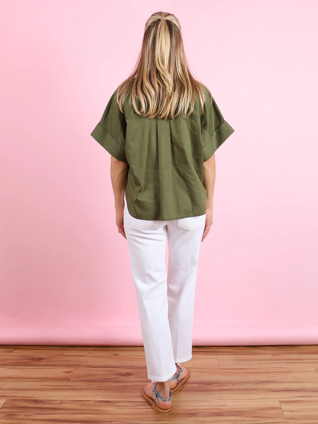 A woman with long blonde hair, partially tied back, faces away from the camera against a pink background wearing the CLOSED Turn-Up Shirt in olive green, paired with white jeans and brown sandals.