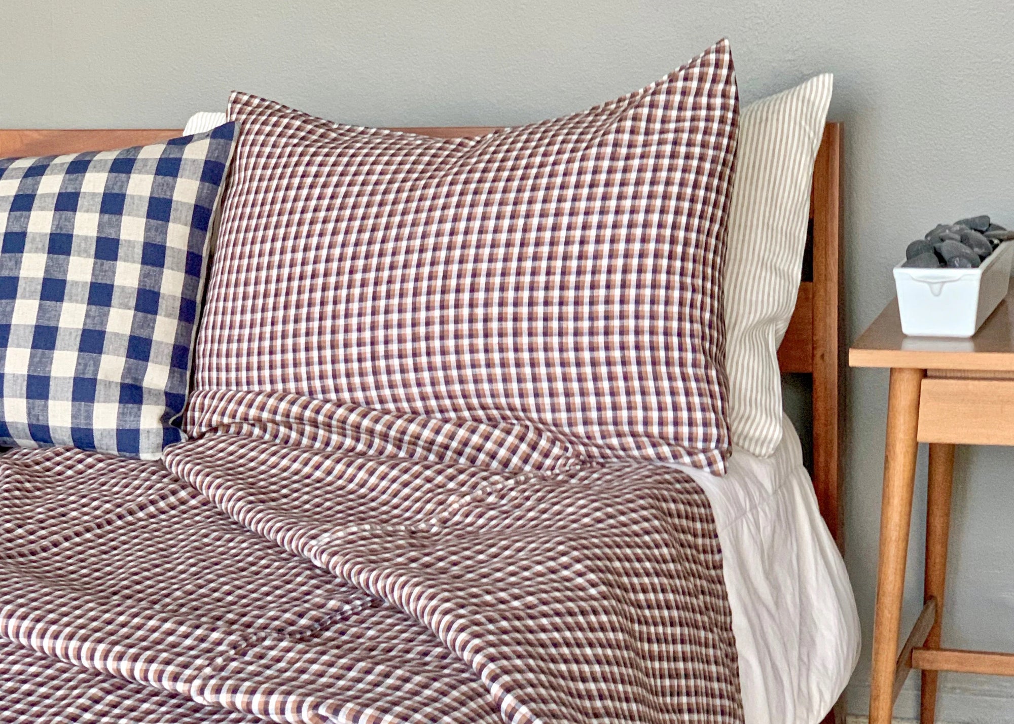 The Hertford King Duvet by Modernplum, featuring brown and white checks, is styled on a neatly made bed with a blue check pillow and nightstand holding a tray of dark stones, set against a light wall.
