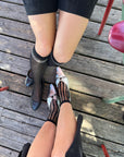Two people sit on wooden decking, legs crossed, wearing black shorts and Unified Sheer Bubble Socks. One pairs the socks with black shoes, the other with silver shoes featuring black stripes. Red chair legs are visible nearby.