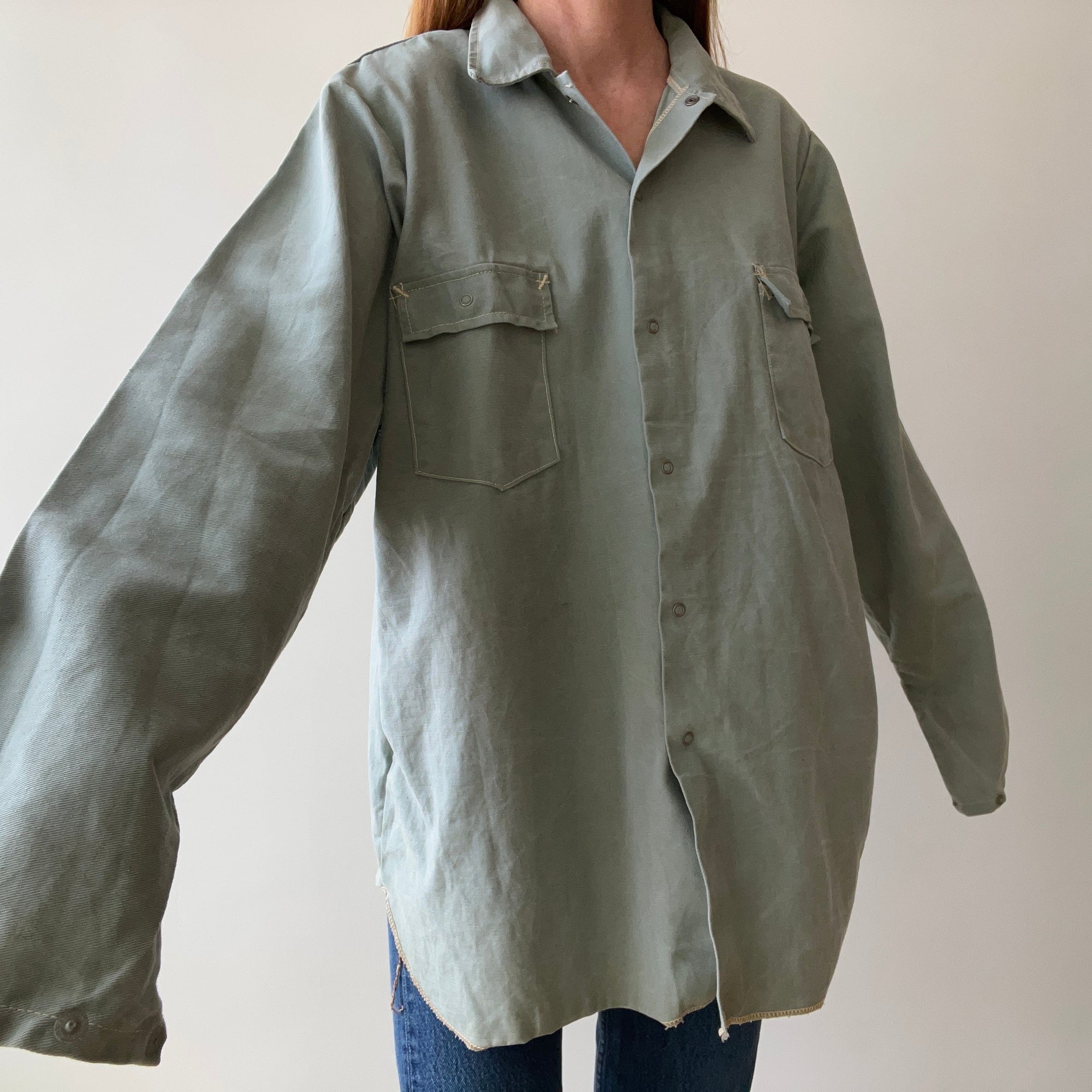 A person wears the Red Vintage Co. 1950s Sage Canvas Workshirt—an oversized, heavyweight sage green button-up with two chest pockets and long, loose sleeves—paired with blue jeans, posed against a plain light background.