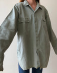 A person wears the Red Vintage Co. 1950s Sage Canvas Workshirt—an oversized, heavyweight sage green button-up with two chest pockets and long, loose sleeves—paired with blue jeans, posed against a plain light background.