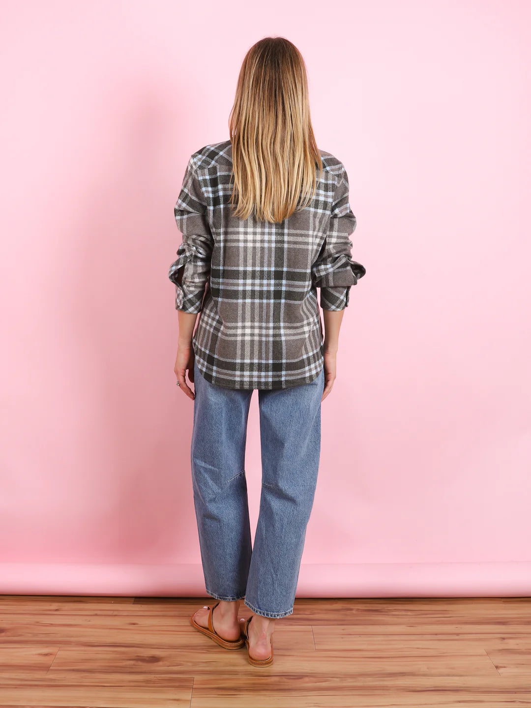 A person with long blonde hair stands facing away, wearing the BCS Group Lumber L/S Shirt, blue jeans, and brown sandals, against a pink background with a wooden floor.