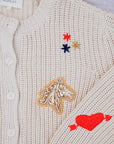 The Collegiate Cardigan by The Great Inc. is a beige knit with white buttons, featuring exquisite hand embroidery of a horse head, a red heart with an arrow, and red, blue, and gold stars.