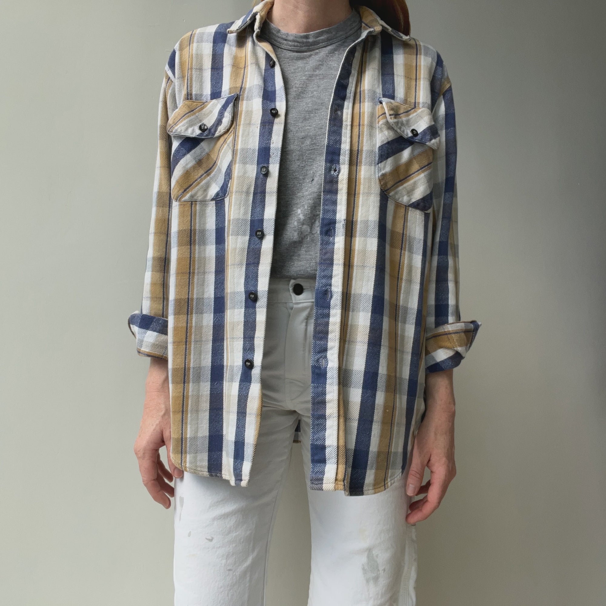 A person dons the 90/2000s Neutrals Navy Flannel from Red Vintage Co., layered over a gray t-shirt to embody classic style. The flannel's rolled-up sleeves and vintage-style chest pockets with flaps add a timeless touch, while white pants complete the ensemble against a simple, light-colored wall backdrop.