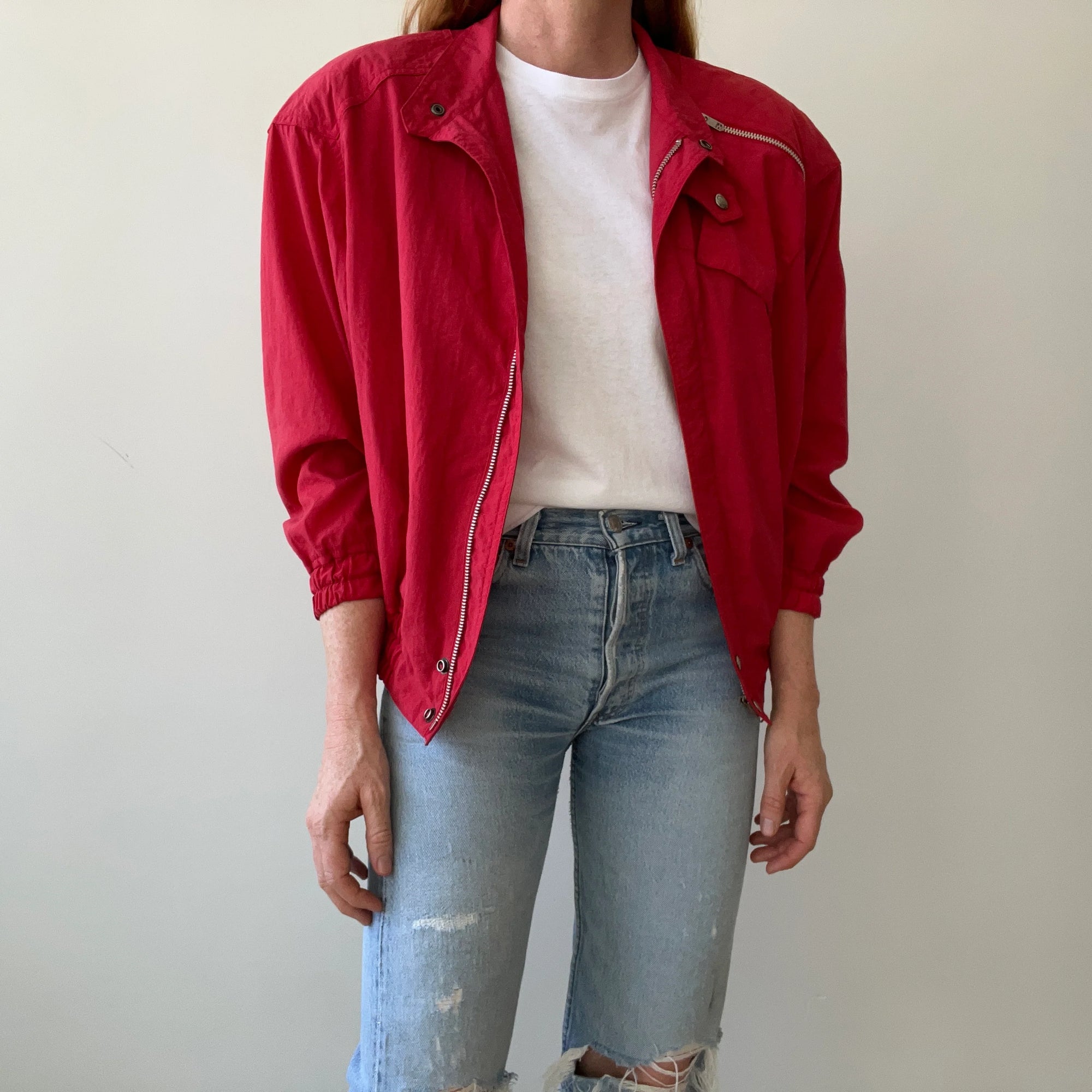A person wearing the 1980s Alpha Cubic Racing Jacket by Red Vintage Co.—a bright red zip-up windbreaker—over a white t-shirt and light blue ripped jeans stands against a plain, light background, with their head not visible in the image.