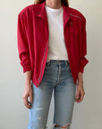 A person wearing the 1980s Alpha Cubic Racing Jacket by Red Vintage Co.—a bright red zip-up windbreaker—over a white t-shirt and light blue ripped jeans stands against a plain, light background, with their head not visible in the image.
