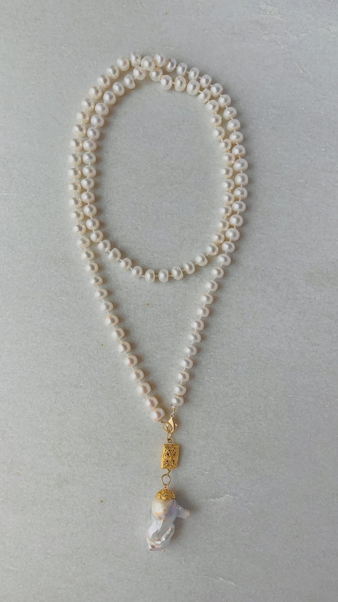 The Mala Ivory with Pearl necklace by Bittersweet Designs is intricately looped into a figure-eight pattern. Suspended at the bottom, akin to prayer beads, is a distinctive, irregularly shaped pearl hanging from a gold-embellished bead, all elegantly displayed against a light grey background.