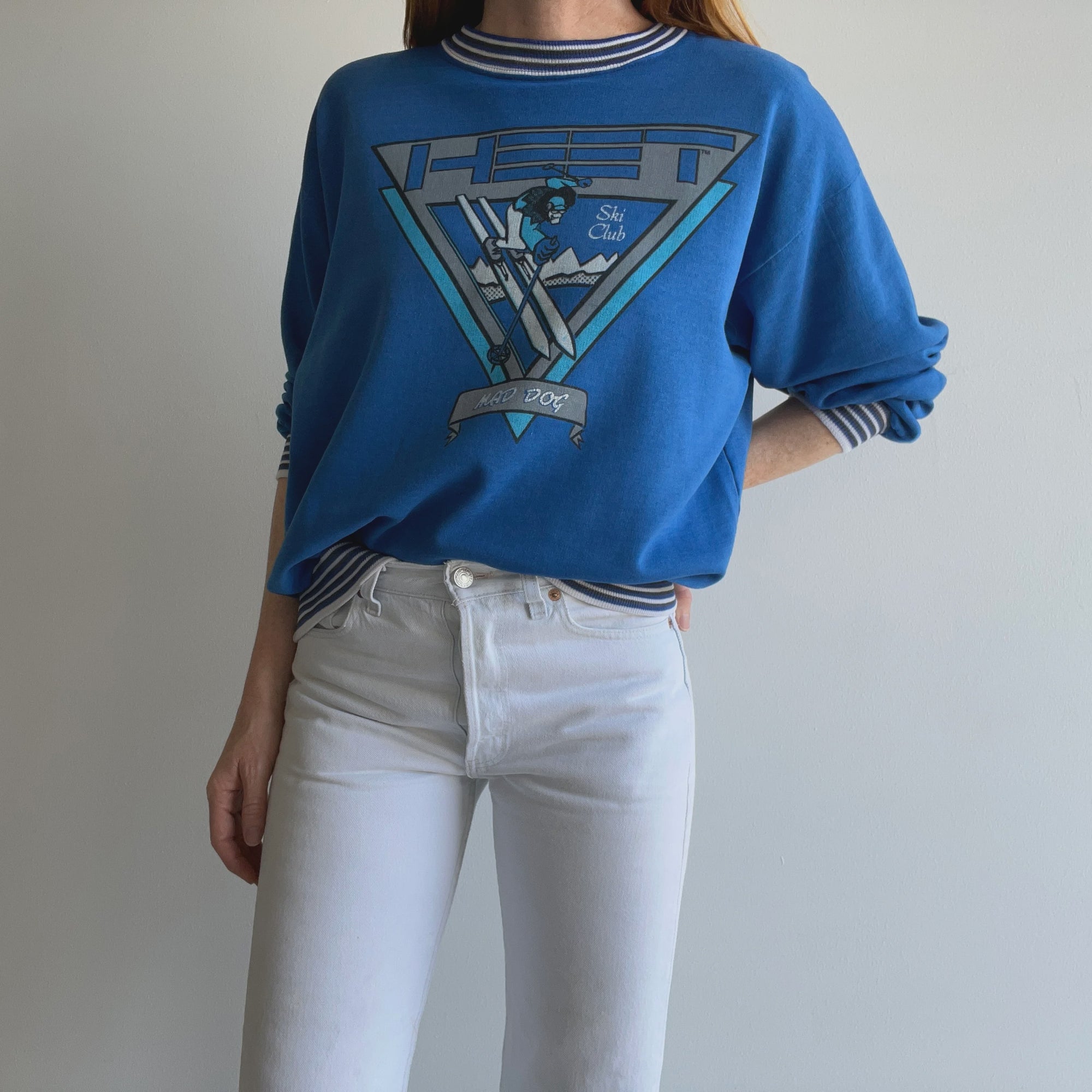 A person wearing an 80s Mad Dog Ski Club Sweatshirt from Red Vintage Co., paired with white high-waisted jeans, stands against a plain white background. The sweatshirt has a soft texture, features a retro ski graphic and striped cuffs, and offers an L/XL fit.