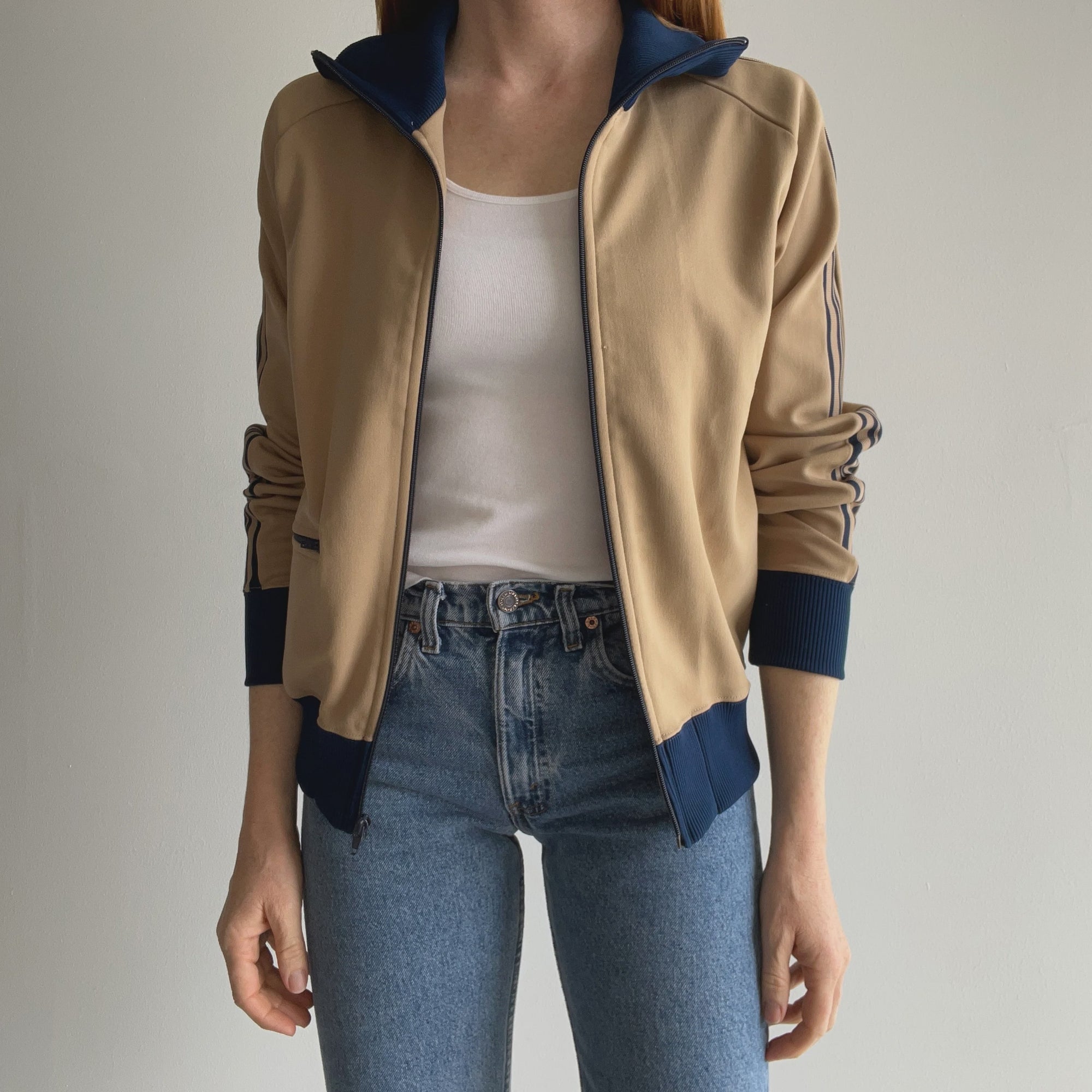 A woman wears the 70s Tracksuit Zip Up Khaki Navy from Red Vintage Co. over a white shirt and blue jeans. Against a plain background, with her hands by her sides, she embodies effortless style.