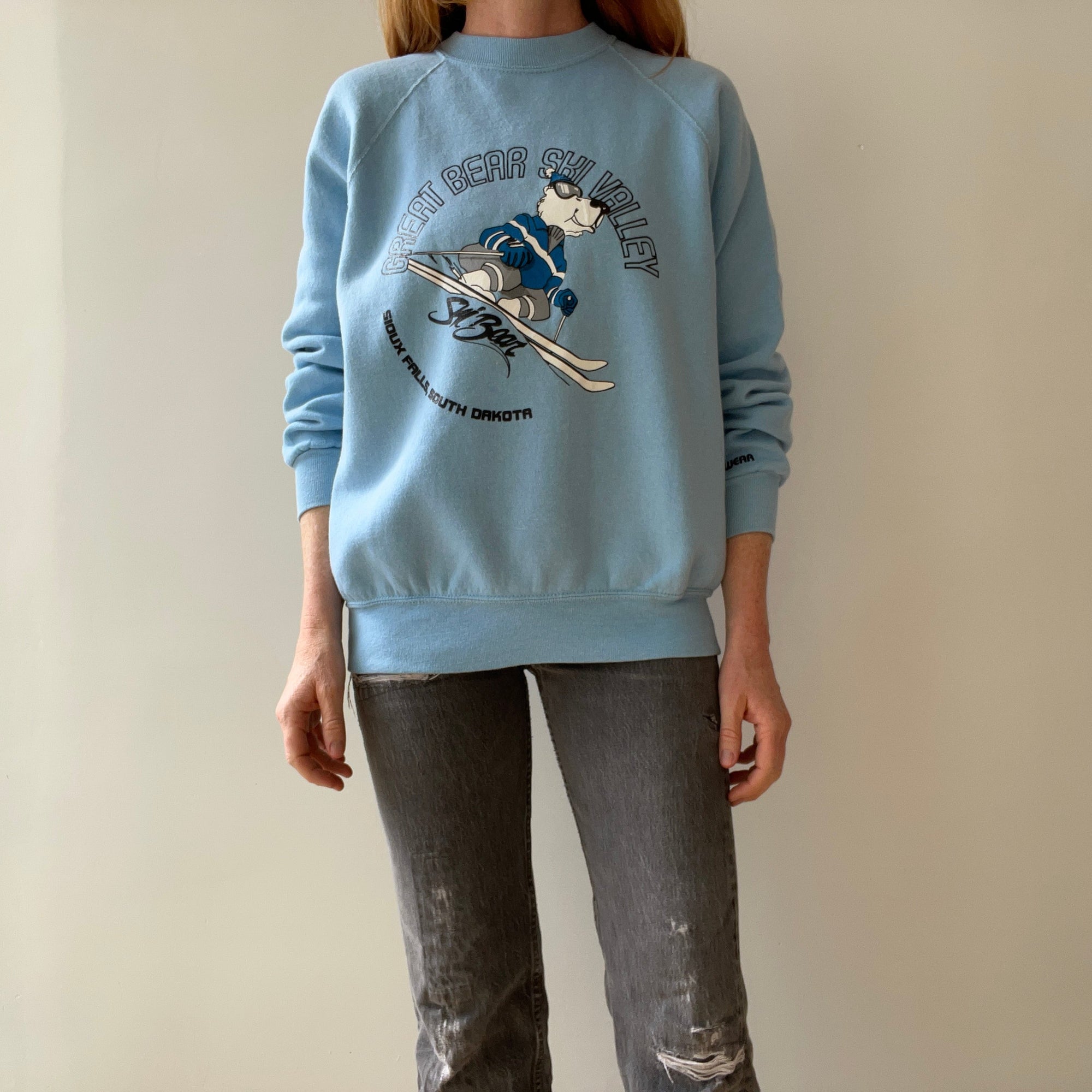 A person wearing the 80s Great Bear Ski Valley Sweatshirt from Red Vintage Co. channels vintage clothing vibes against a plain backdrop. The blue sweatshirt, adorned with a bear skiing graphic and the text "Mount Bear Ski Valley, South Dakota," pairs perfectly with gray jeans for an effortlessly cool 80s fashion look.