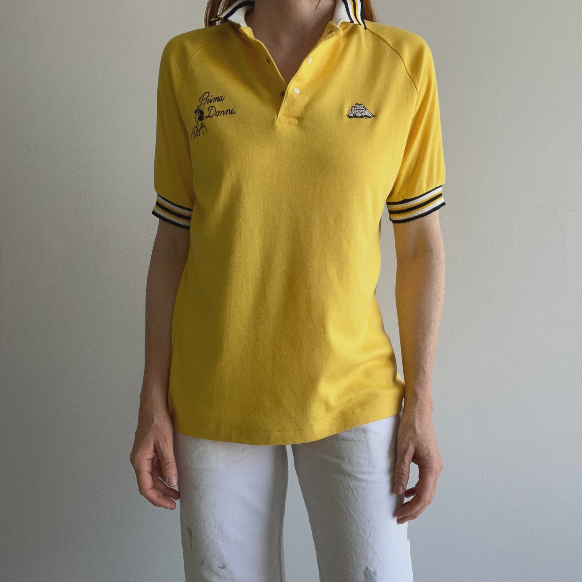 Donning the 70s Prima Donna Jersey knit Polo Shirt by Red Vintage Co., featuring a bright yellow hue with a black and white striped collar and sleeves, embroidered text, and a small logo, the person exudes a classic vintage clothing flair. This look is perfectly balanced with white pants against a plain light wall.