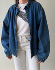 A person models the Red Vintage Co. 90s RL Denim Dad Jacket with plaid details, layered over a white graphic tee and high-waisted white jeans, standing against a plain light background. Their head is not shown.