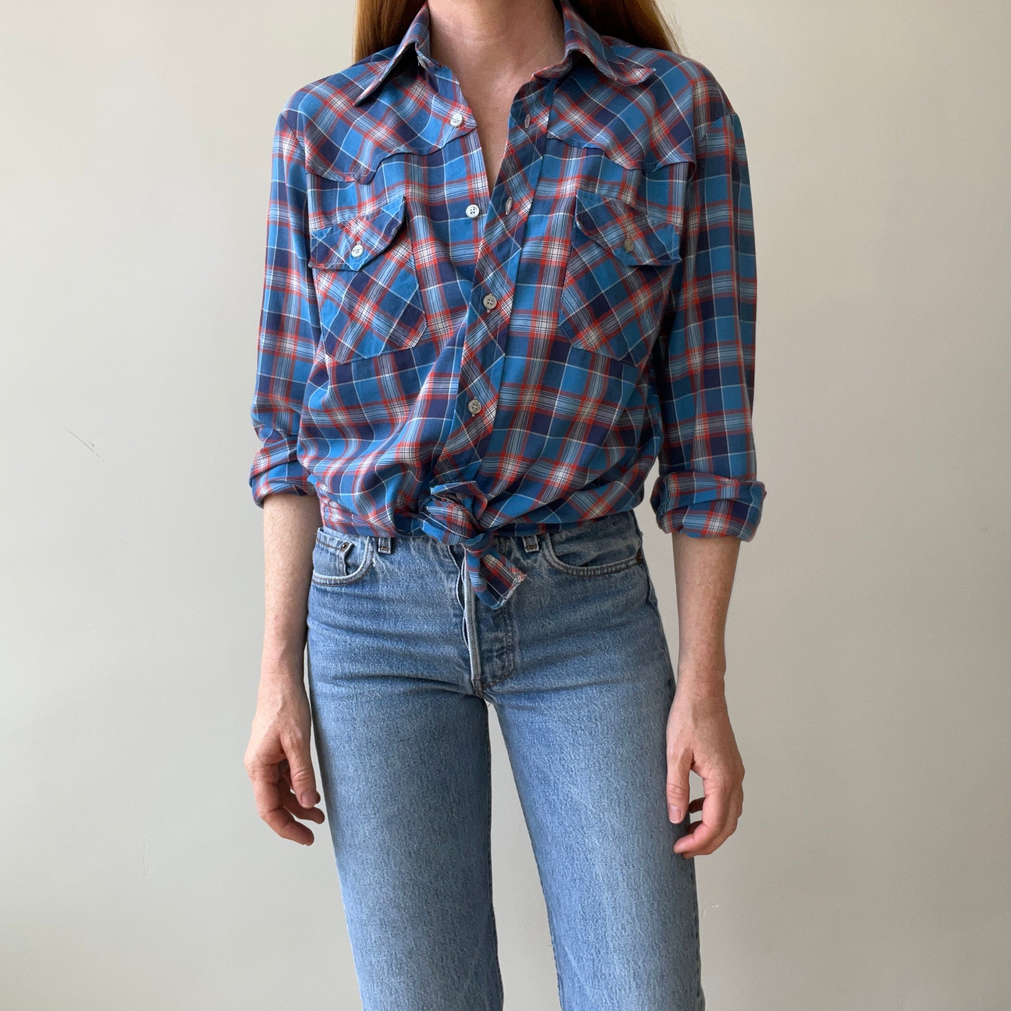 A person wears the 1970s Plaid Western shirt by Red Vintage Co., tied at the waist, with loose-fit, ultra-lightweight high-waisted light blue jeans, standing against a plain light-colored background.
