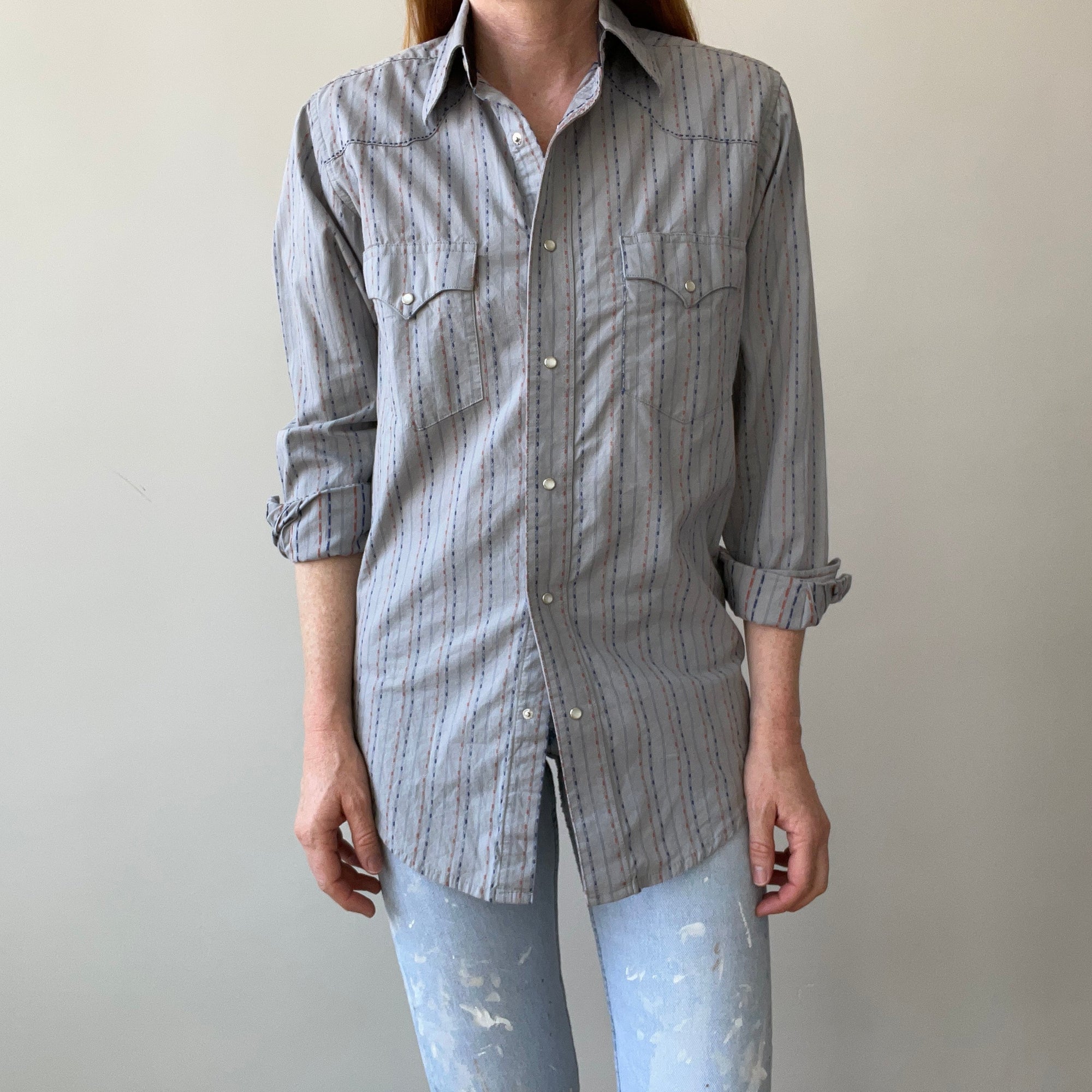 A person models the 80s Wrangler Button Up from Red Vintage Co.—a sun faded, pearl snap shirt with thin vertical stripes—paired with light blue distressed jeans, standing against a plain light background. Face not visible.