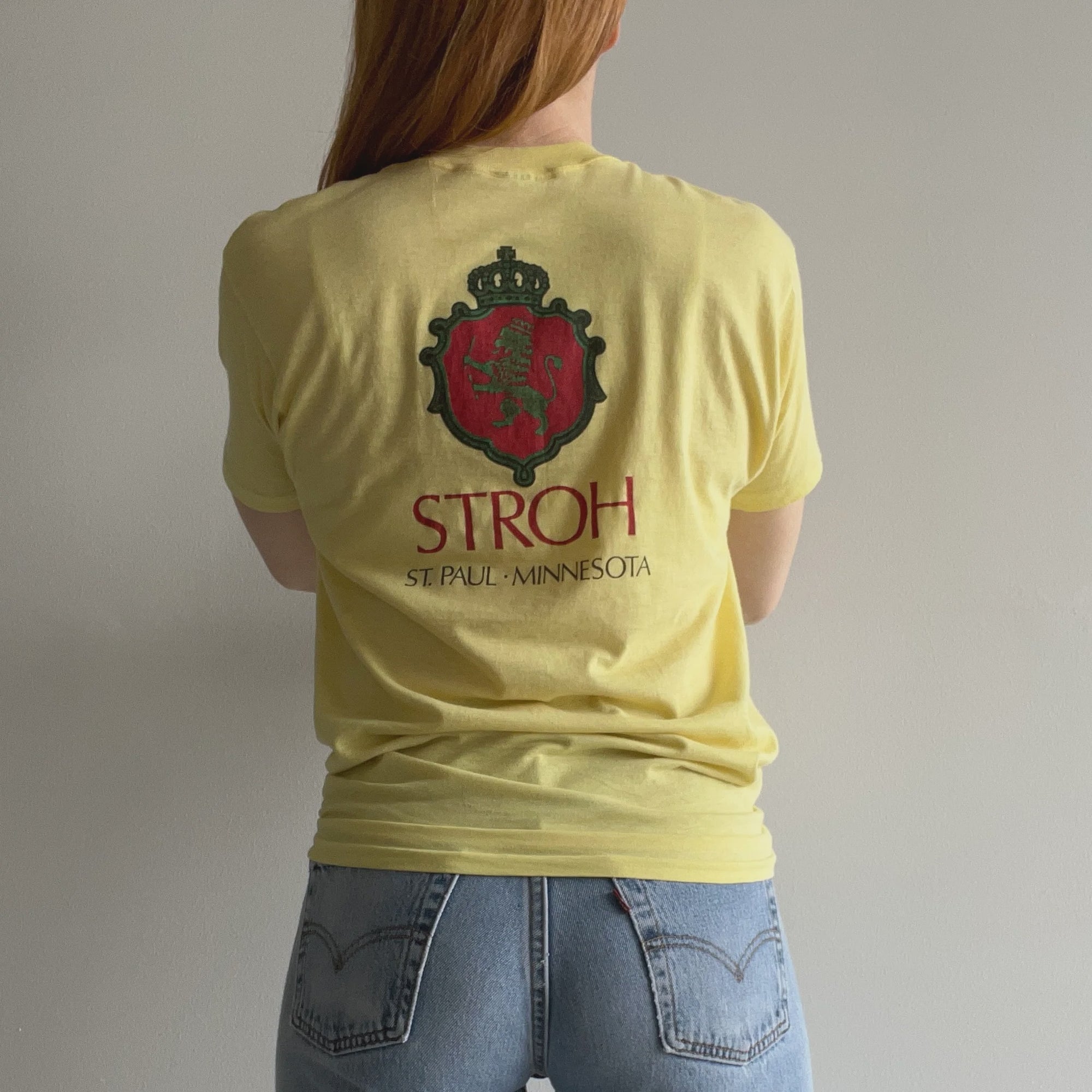 Wearing the 80s Stroh St. Paul MN Beer Tee from Red Vintage Co., a person with long hair stands against a plain wall, showcasing the vintage clothing charm of the yellow T-shirt adorned with a red and black crest and the words "STROH ST. PAUL - MINNESOTA." Complemented by light blue jeans, their look captures the essence of midwestern brew roots.