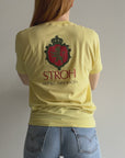 Wearing the 80s Stroh St. Paul MN Beer Tee from Red Vintage Co., a person with long hair stands against a plain wall, showcasing the vintage clothing charm of the yellow T-shirt adorned with a red and black crest and the words "STROH ST. PAUL - MINNESOTA." Complemented by light blue jeans, their look captures the essence of midwestern brew roots.