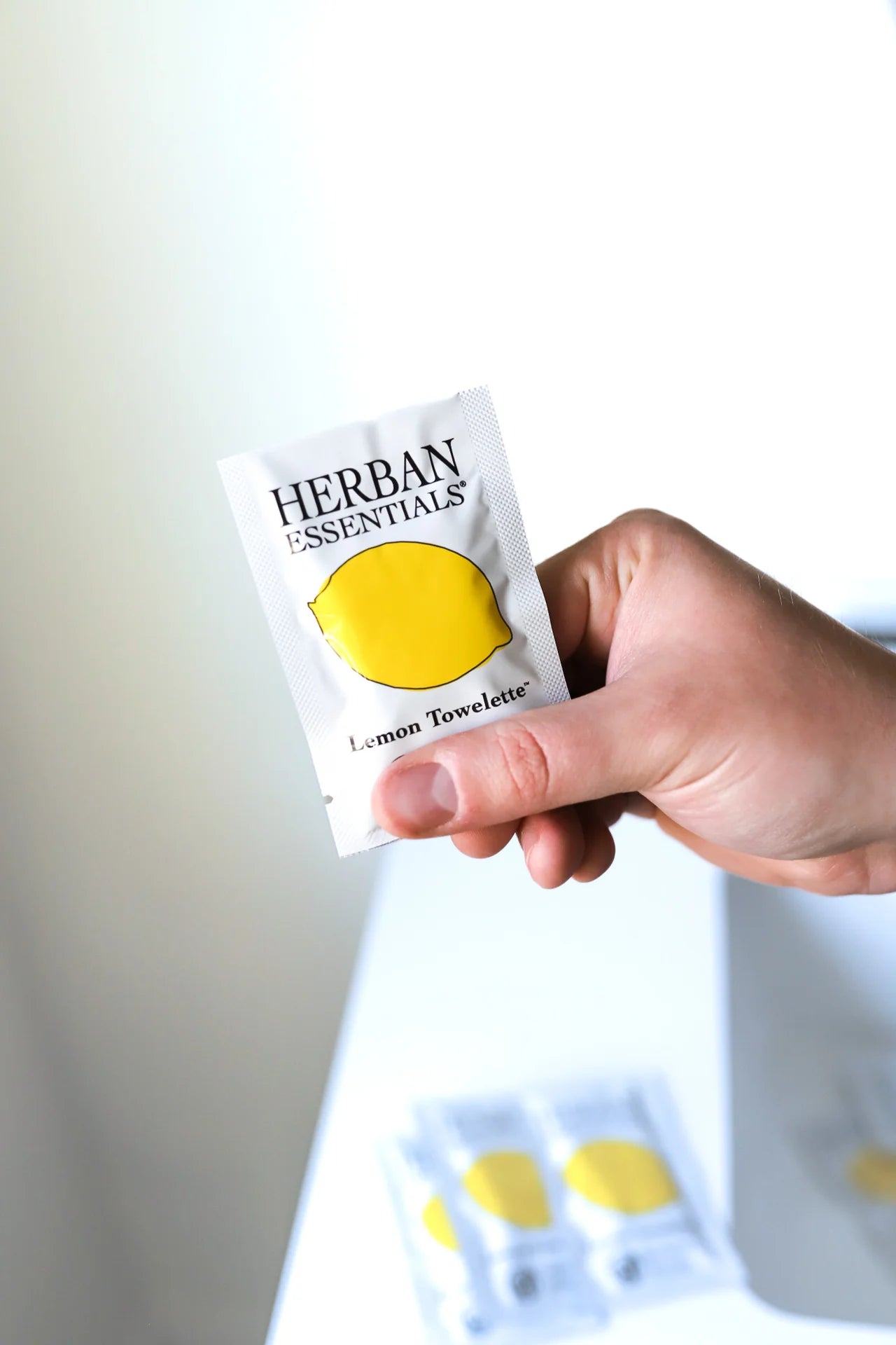 A hand holds a Herban Essentials Essential Oil Towelettes packet featuring lemon imagery, emphasizing its refreshing lemon scent; two more towelette packets are blurred in the background on a white surface.